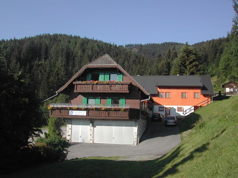 Tradičný hostinec s drevenými balkónmi a zelenými okenicami pred zalesneným kopcom.