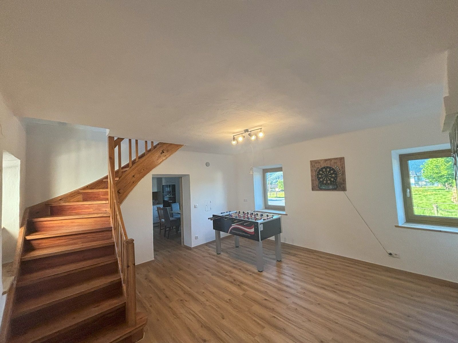 Spoločenská miestnosť so stolným futbalom, šípkami na stene, dreveným schodiskom a dvoma oknami.