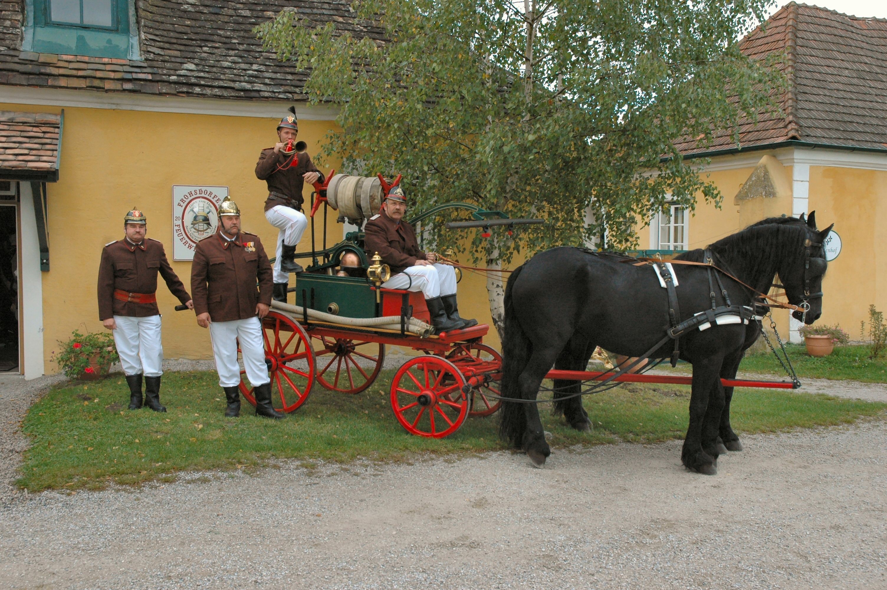 Historické hasičské auto s koňom a hasičmi v uniformách pred žltou budovou.