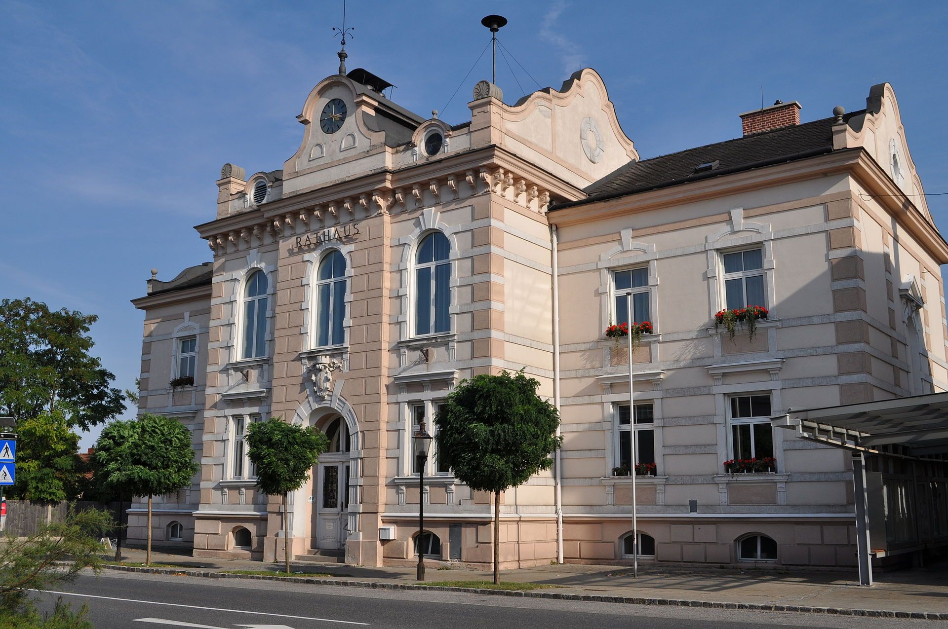 Radnica mesta Felixdorf, historická budova s hodinovou vežou a ozdobnými oknami, obklopená stromami a ulicou v popredí.