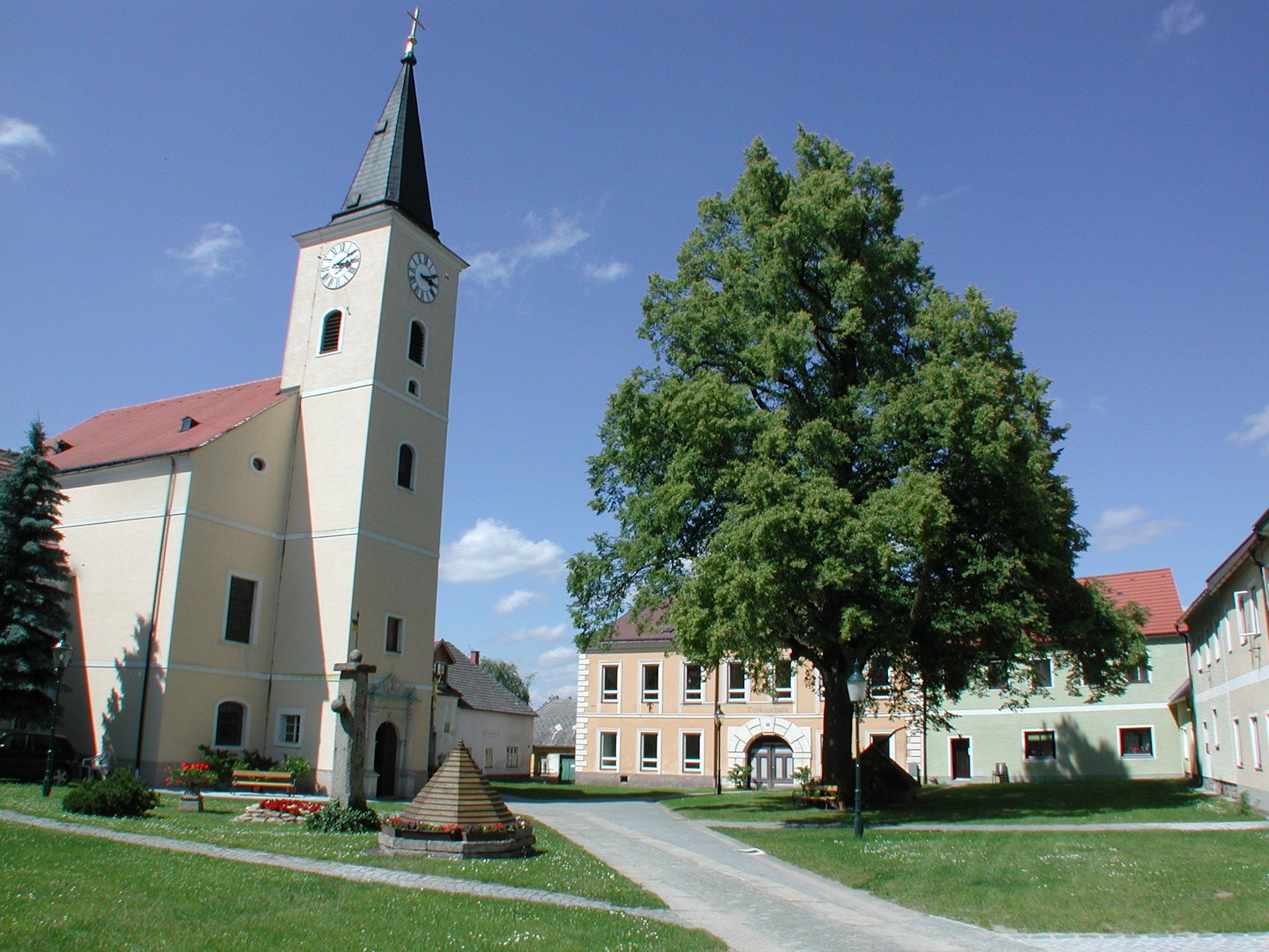 Kostol s vežou a hodinami na dedinskom námestí so stromami a budovami.