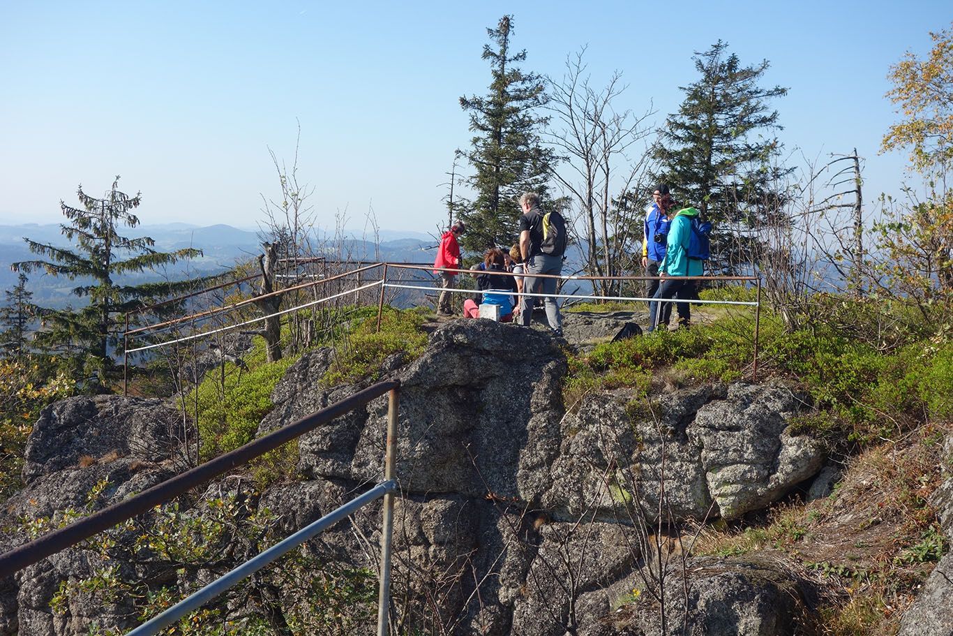 Skupina turistov na skalnatom vyhliadkovom mieste so zábradlím, obklopená stromami a horami v pozadí.