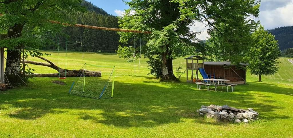 Detské ihrisko na zelenej lúke s hojdačkami, lezeckou konštrukciou, šmýkačkou a ohniskom. V pozadí sú vidieť stromy a kopce.