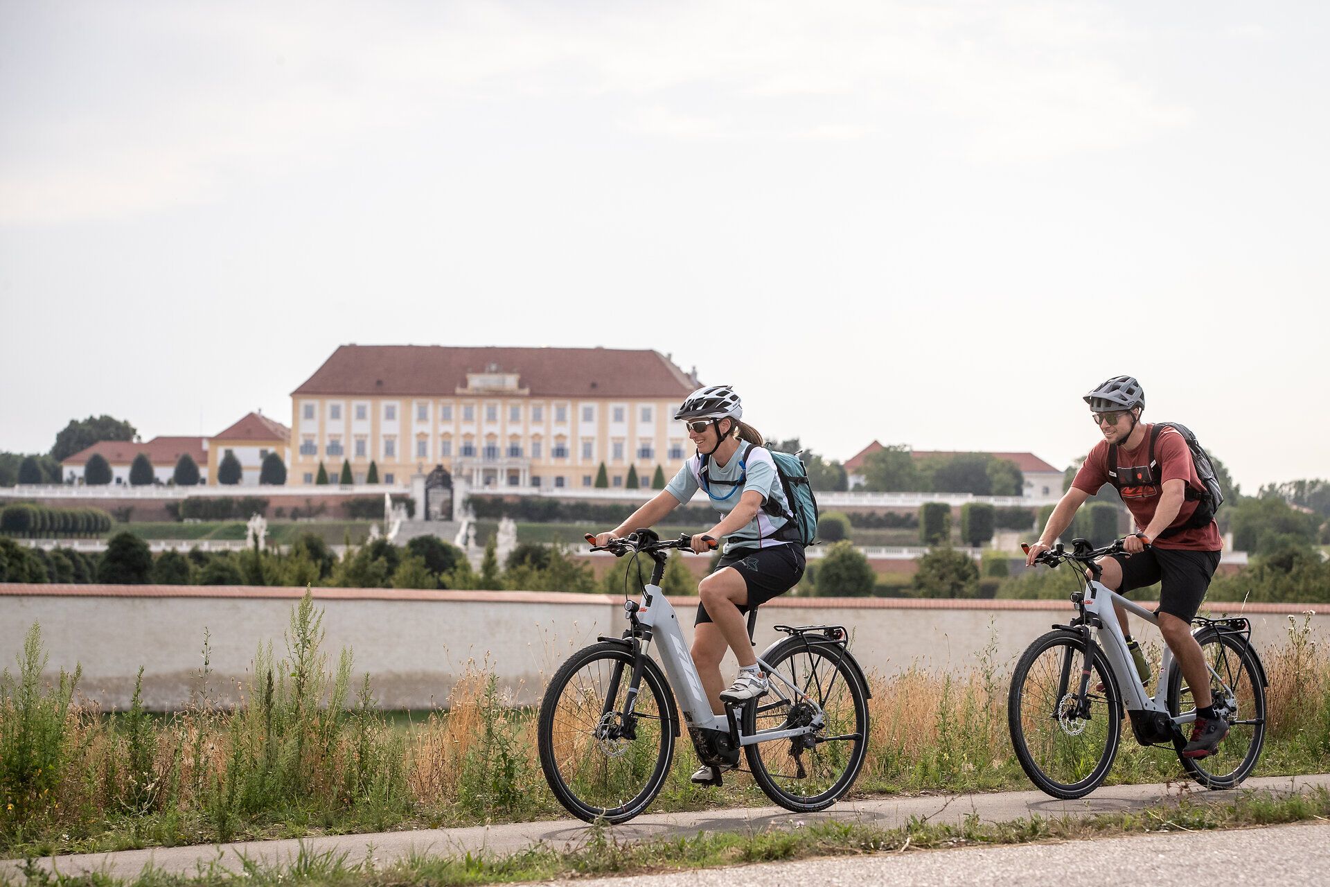 Dvaja cyklisti si vychutnávajú čerstvý vzduch a malebnú krajinu pozdĺž tichého kanála. Zvlnené kopce v pozadí a bujná vegetácia vytvárajú príjemnú atmosféru pre milovníkov pohybu v prírode. Toto je leto v tej najlepšej podobe.