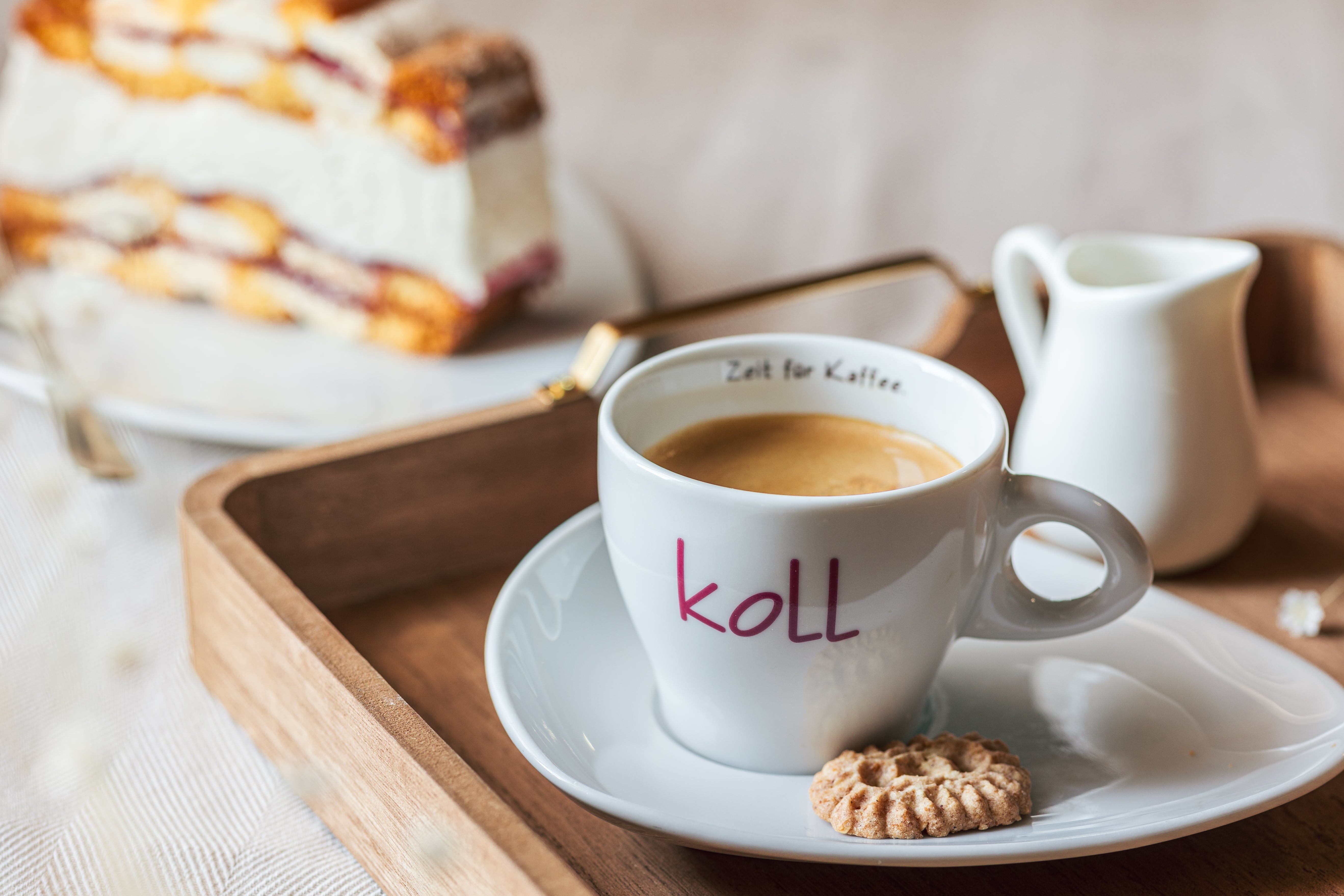 Šálka kávy so sušienkou na podnose, v pozadí kúsok koláča.