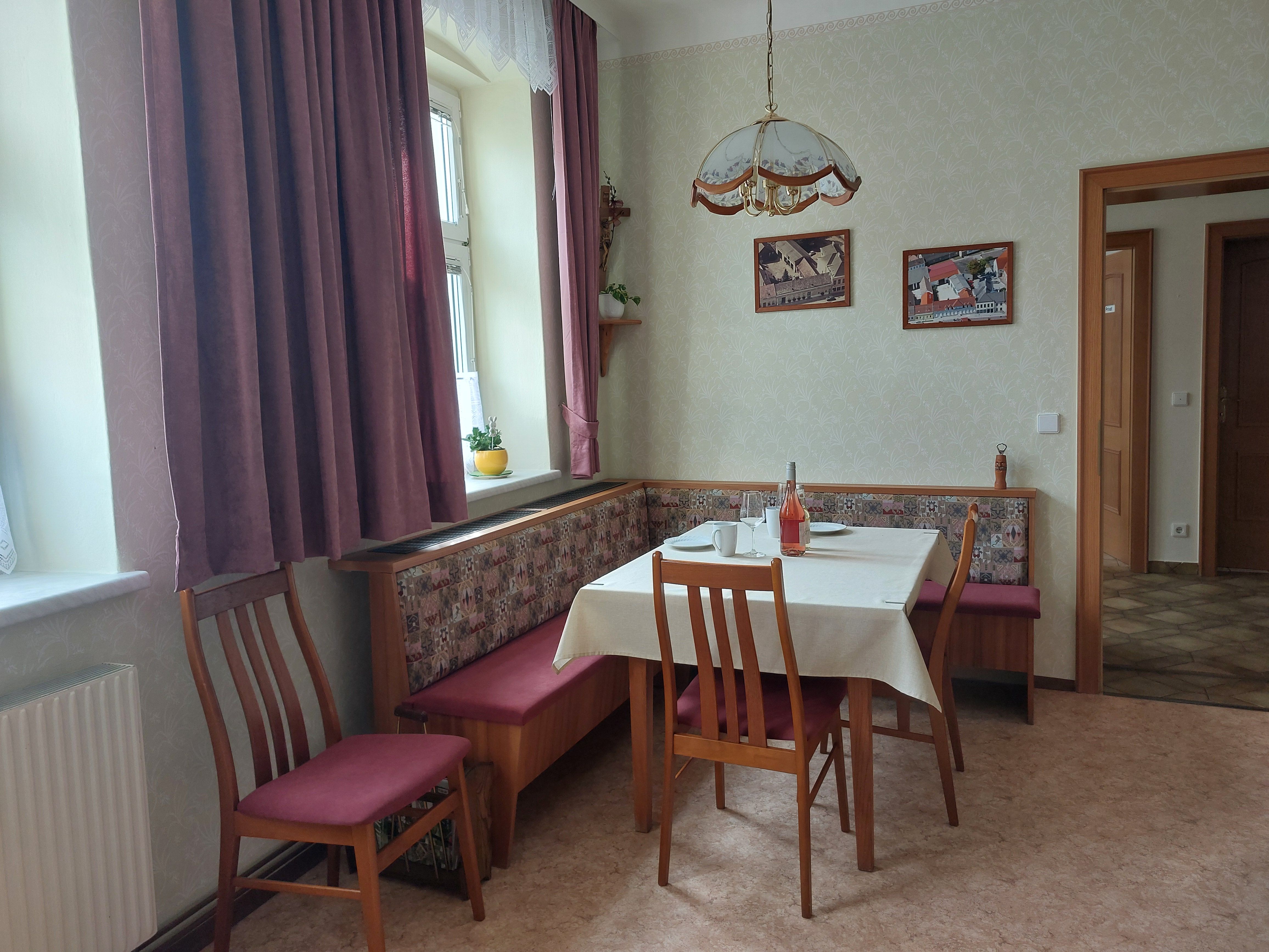 Útulná kuchyňa s jedálenským stolom, stoličkami a rohovou lavicou. Okno so závesmi, obrazy na stene.