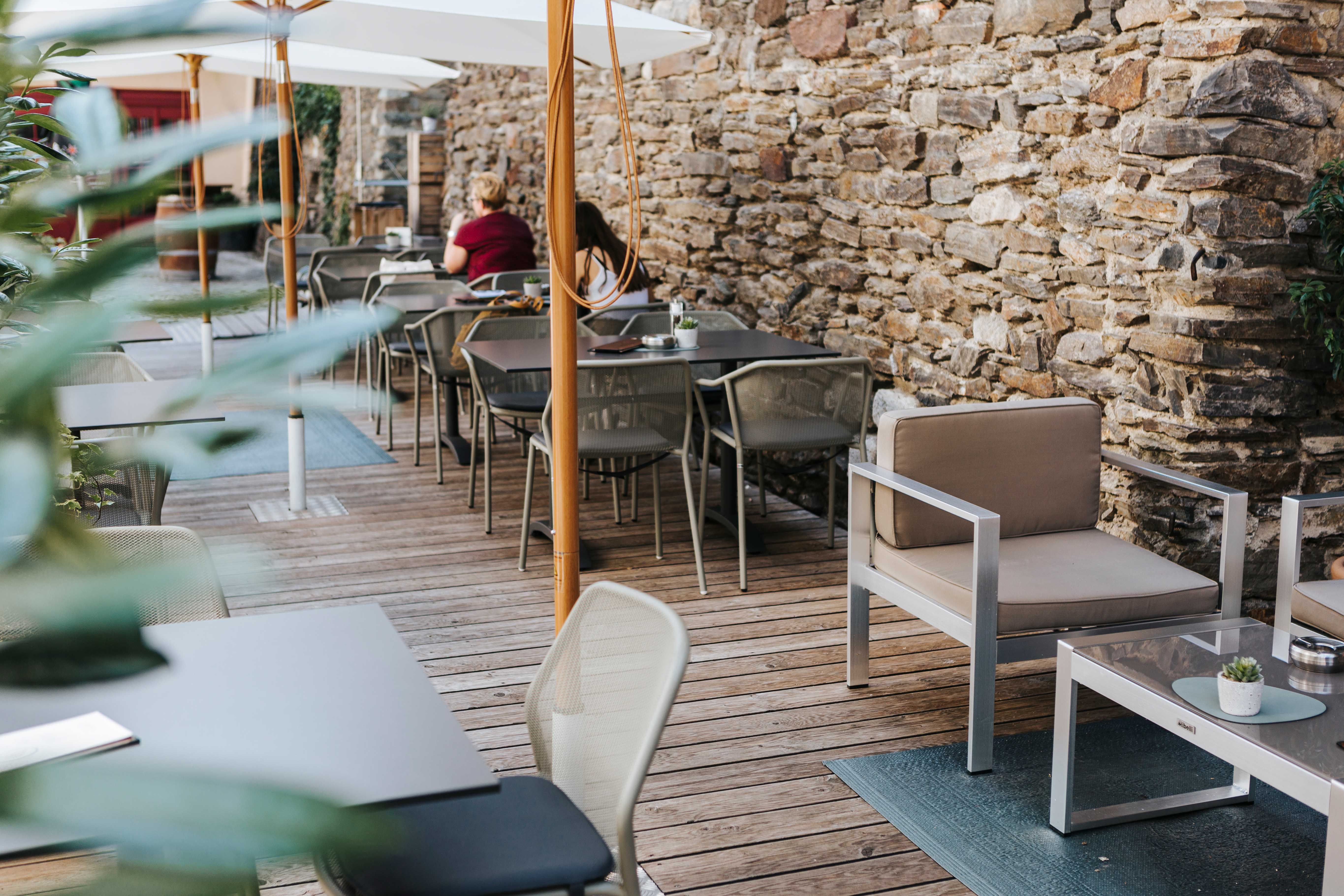 Útulný vonkajší priestor kaviarne s drevenou podlahou, moderným nábytkom a kamennou stenou.