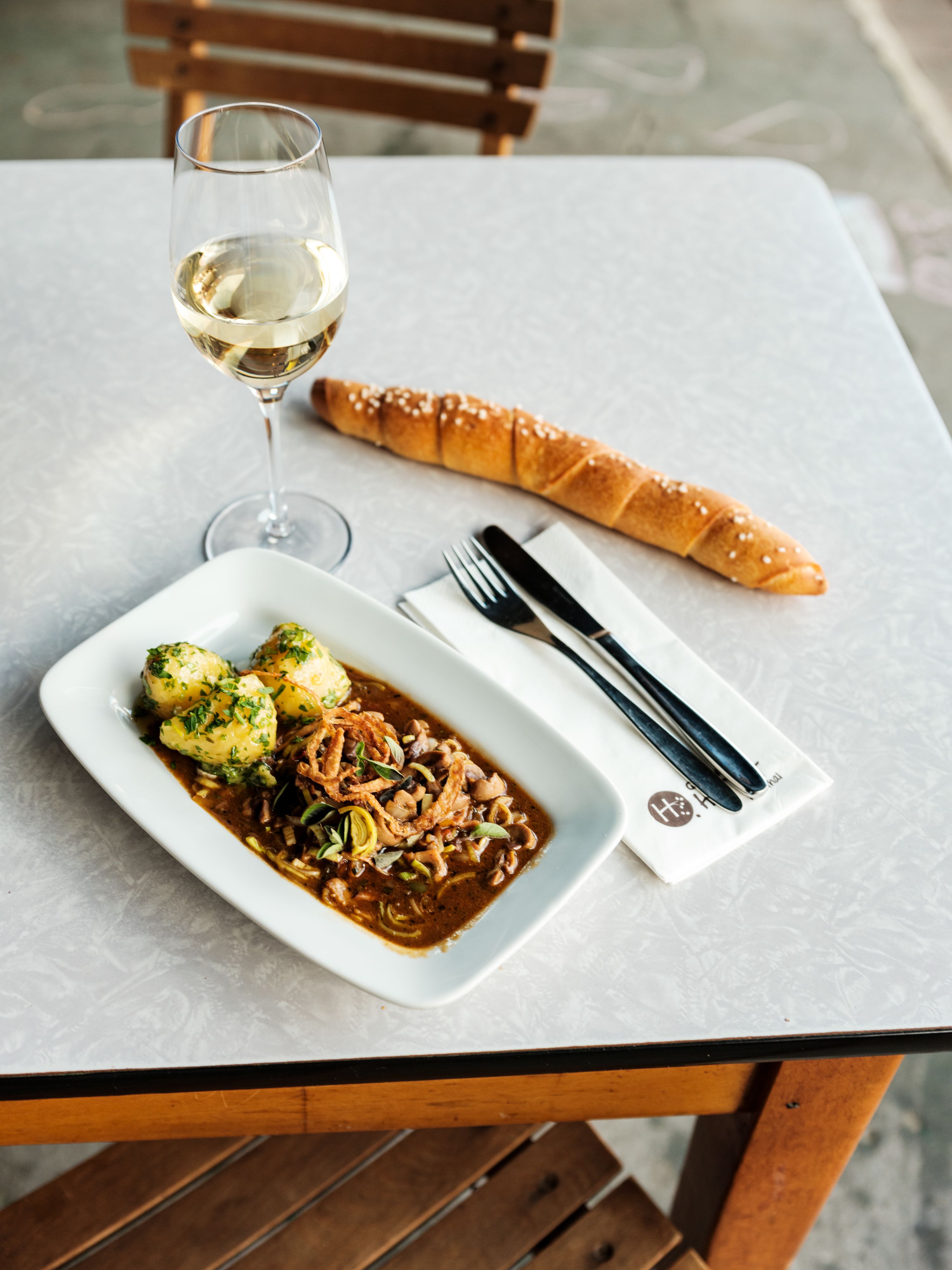 Stôl s pohárom bieleho vína, tanier s mäsovým pokrmom a knedľou, dlhá žemľa a príbor na obrúsku.