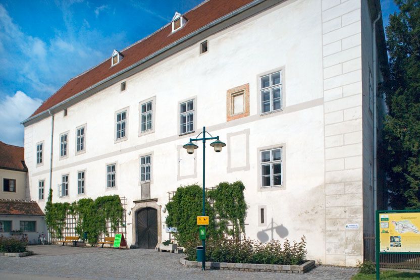 Zámok Traismauer s bielou fasádou a červenými škridlami, obklopený rastlinami a vydláždeným predpolím.
