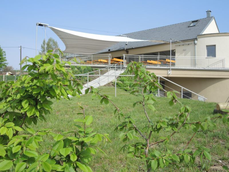 Vínna taverna s terasou a markízou, obklopená zelenou krajinou.