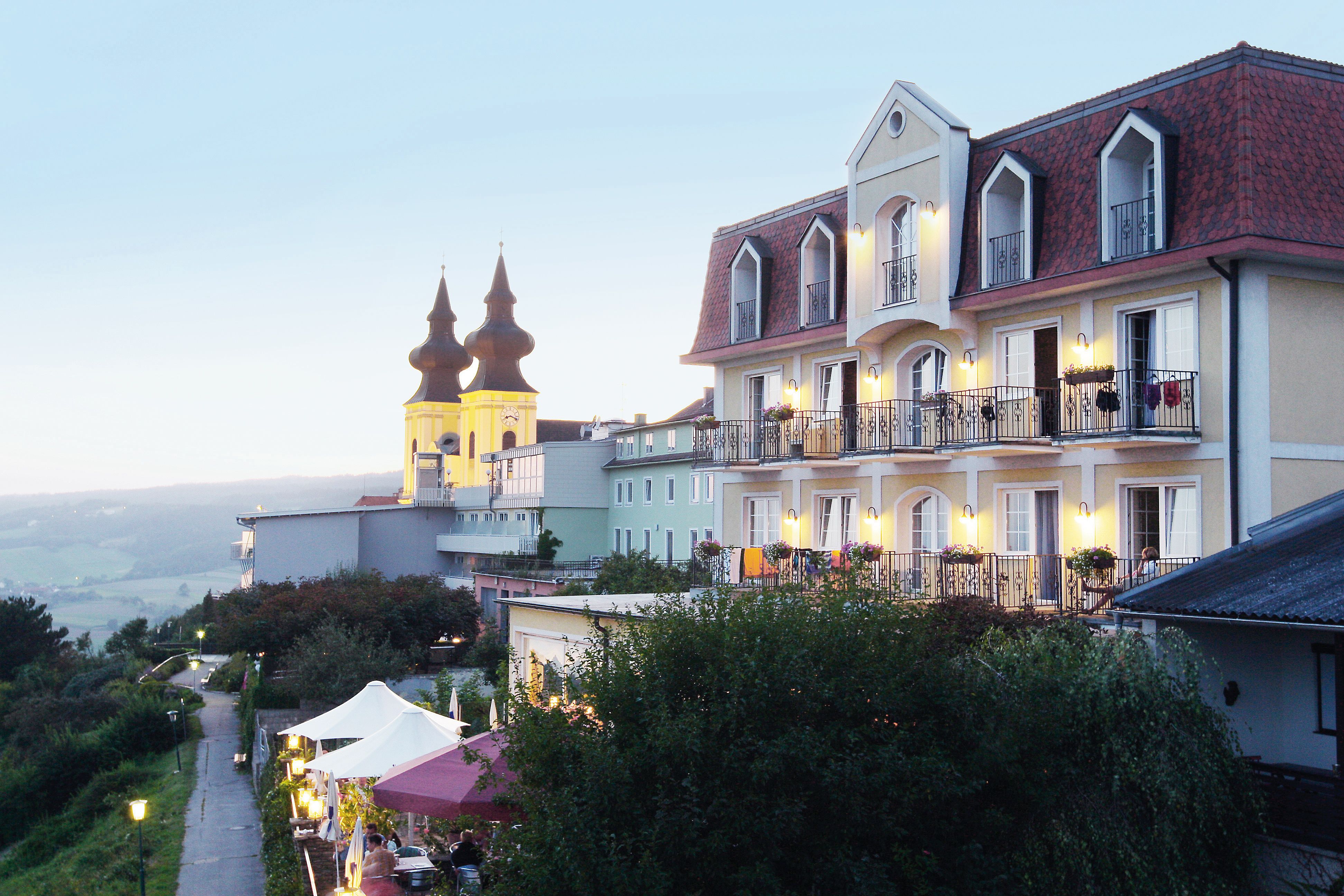 Hotel s balkónmi a kostolom v pozadí za súmraku.