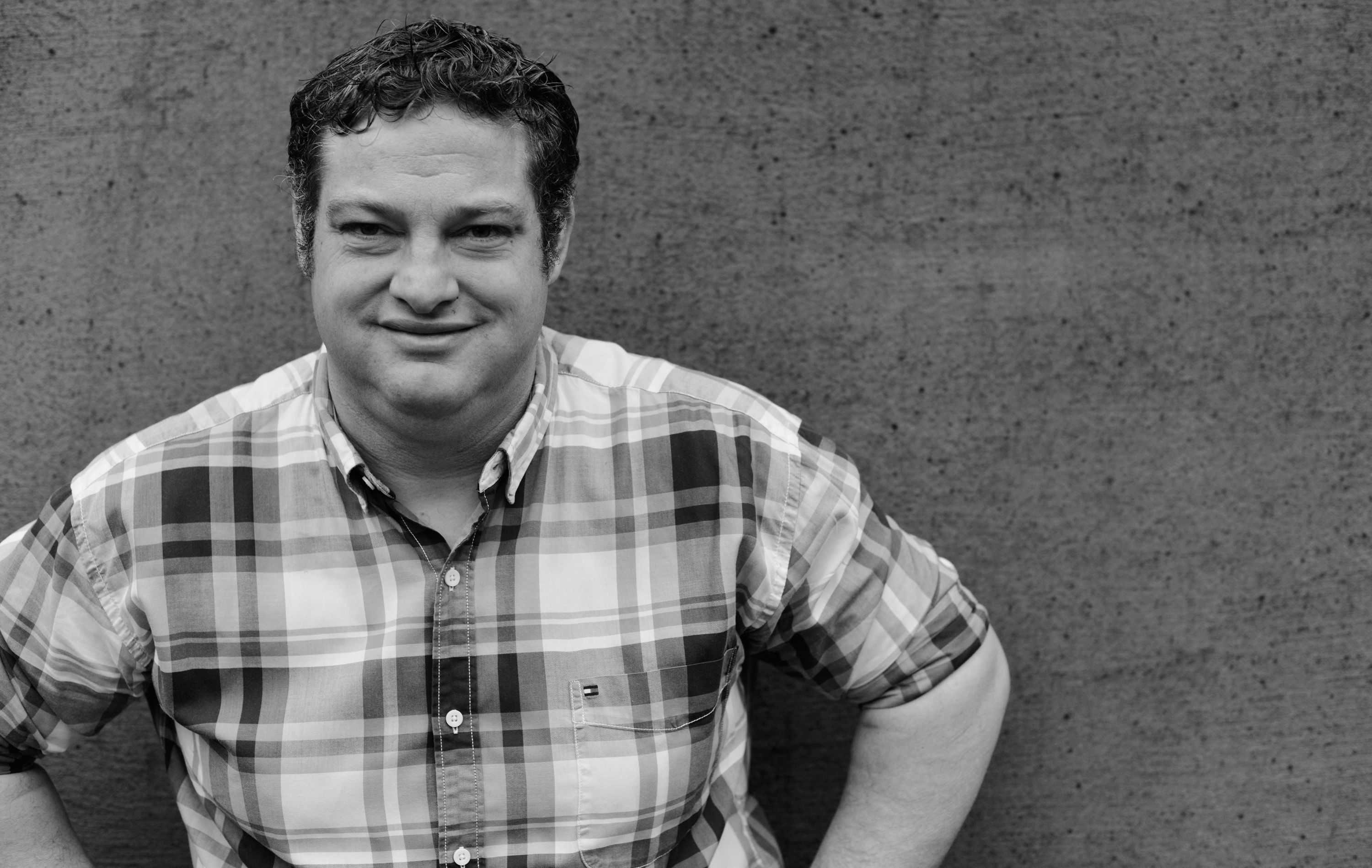Čiernobiela fotografia muža v kockovanej košeli pred betónovým múrom.