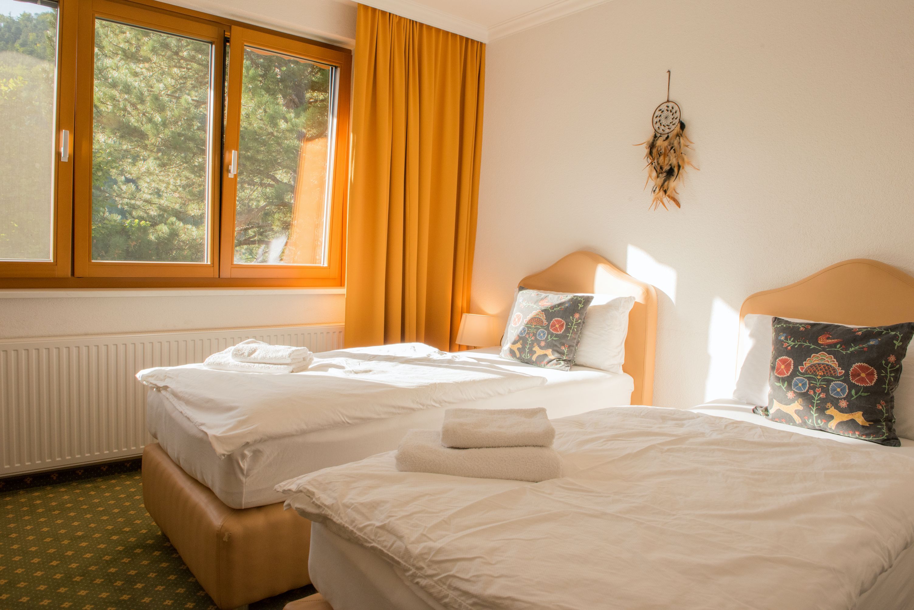 Svetlá dvojlôžková izba s dvoma samostatnými posteľami, žltými závesmi a oknom s výhľadom na zeleň.