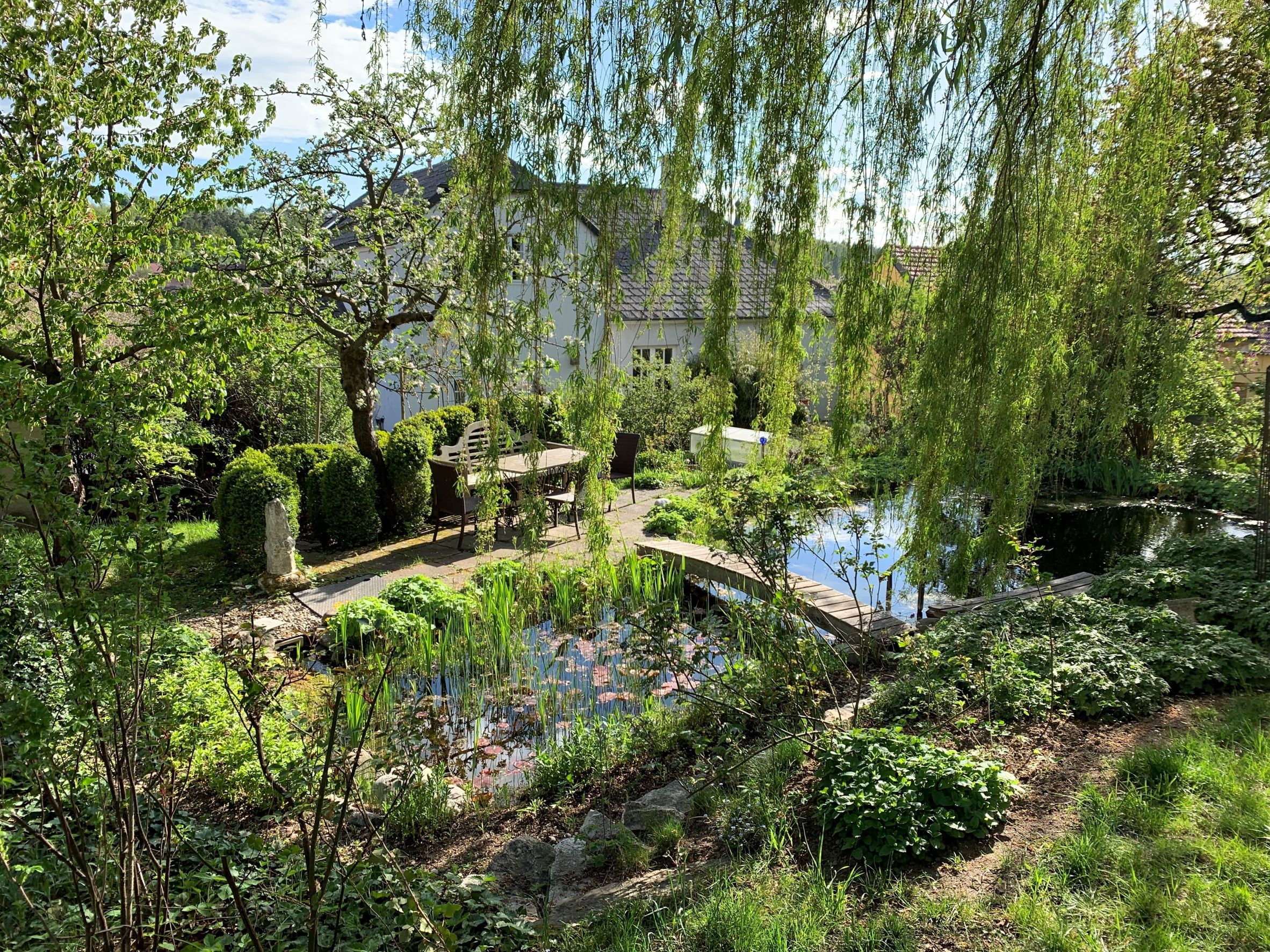 Idylická záhrada s jazierkom, stromami a stolom a stoličkami pod holým nebom.