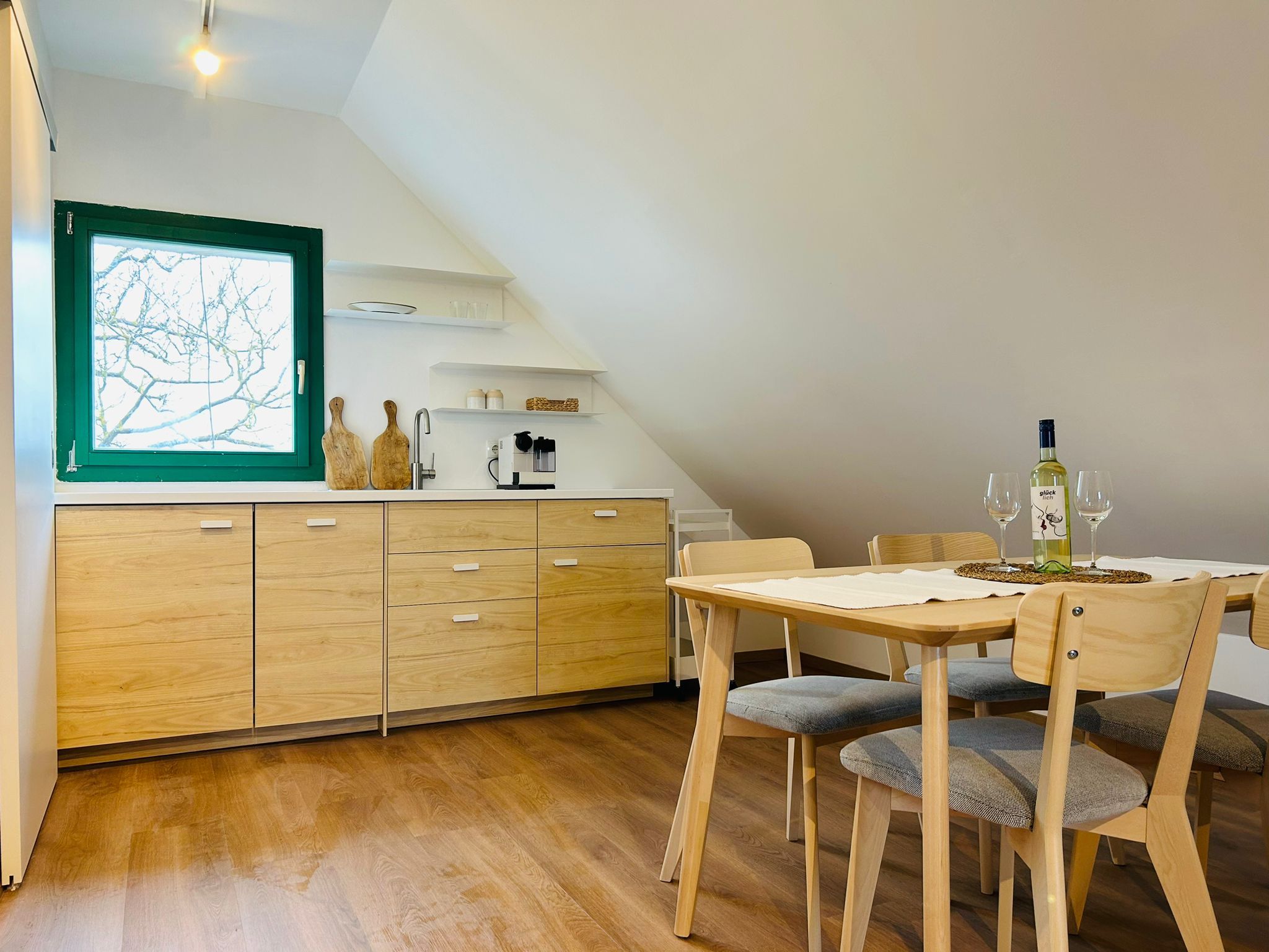 Moderná kuchyňa s drevenými skrinkami, jedálenský stôl a stoličky, okno so zeleným rámom.