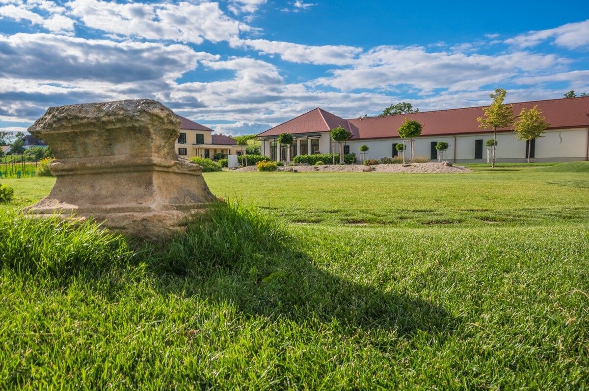 Vinárstvo s trávnikom a antickým kameňom v popredí.