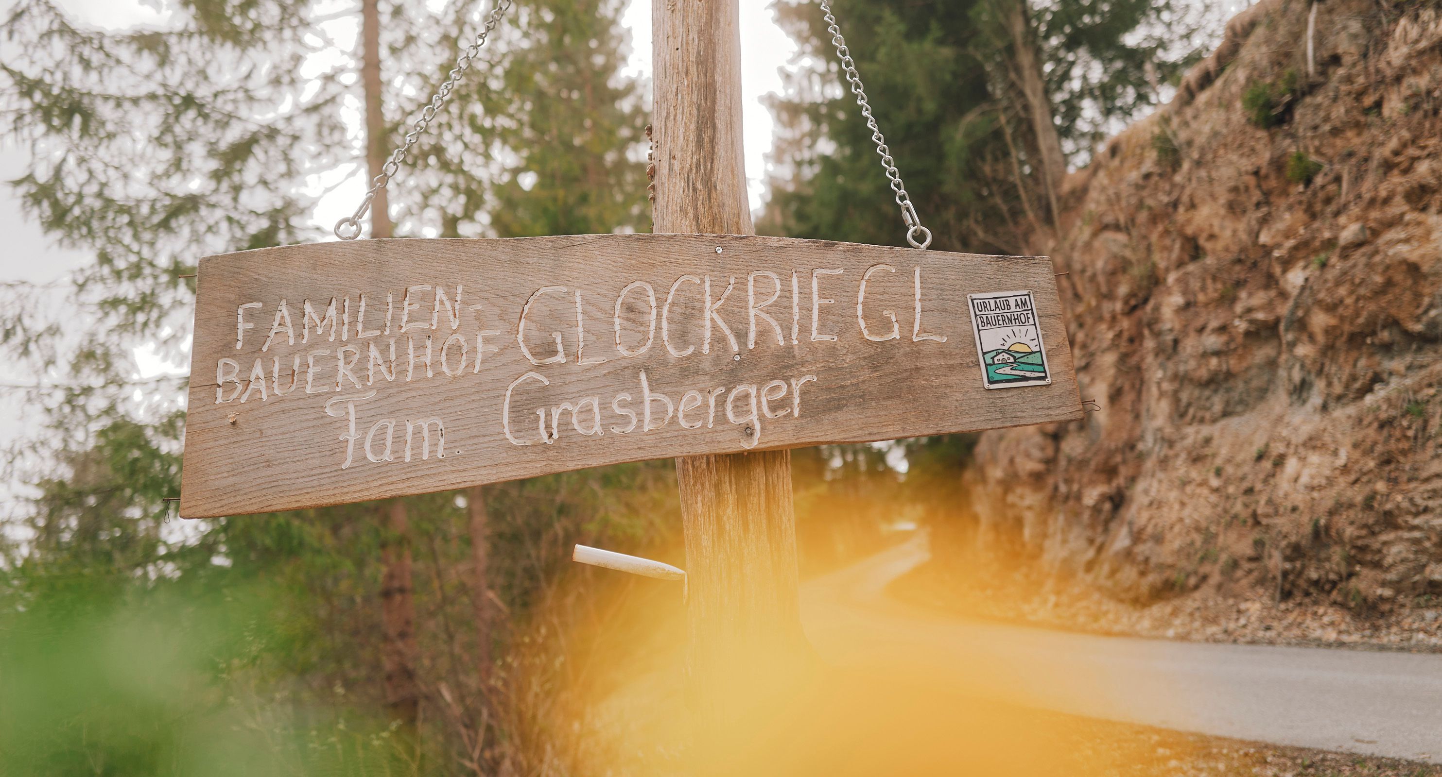 Drevená tabuľa s nápisom "Familienbauernhof Glockriegl Fam. Grasberger" na lesnej ceste.