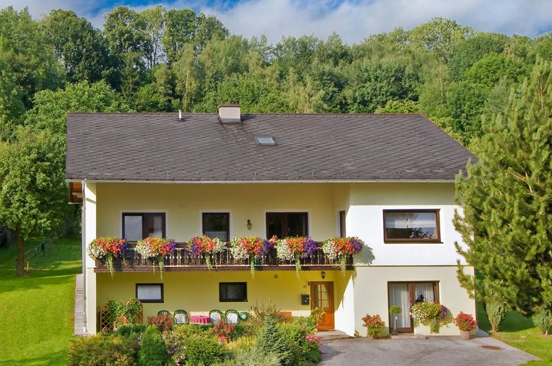 Dvojpodlažný žltý dom s balkónom a kvetinami, obklopený stromami a záhradou.