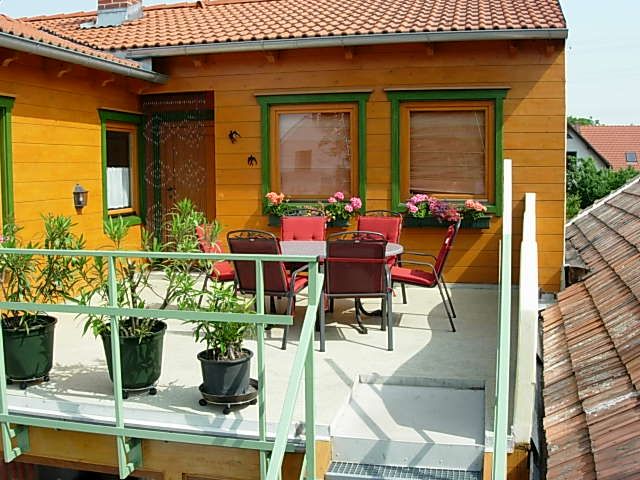 Balkón so stolom, stoličkami a rastlinami, obklopený domom s oranžovou fasádou a zelenými okennými rámami.