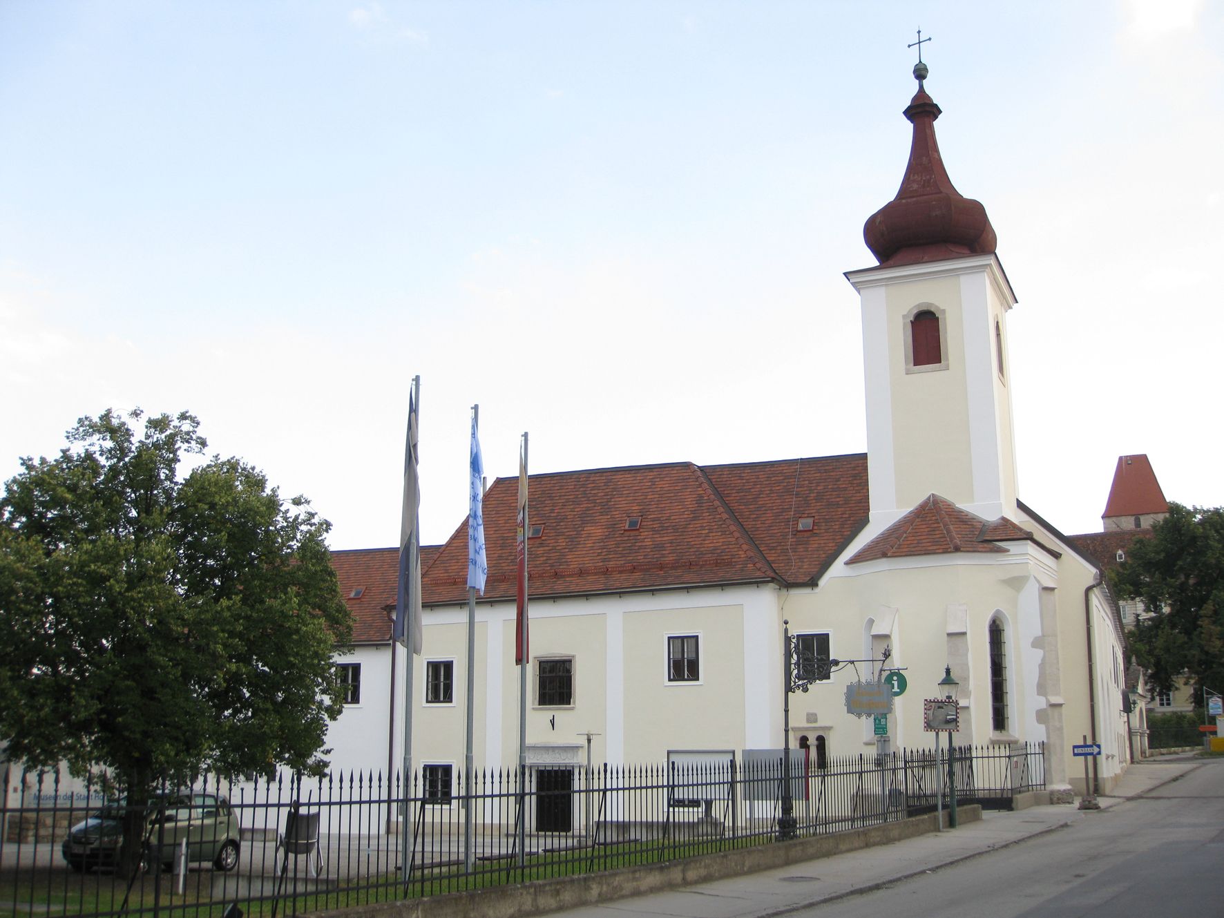 Budova s kostolnou vežou a červenými škridlami, obklopená plotom a stromami.