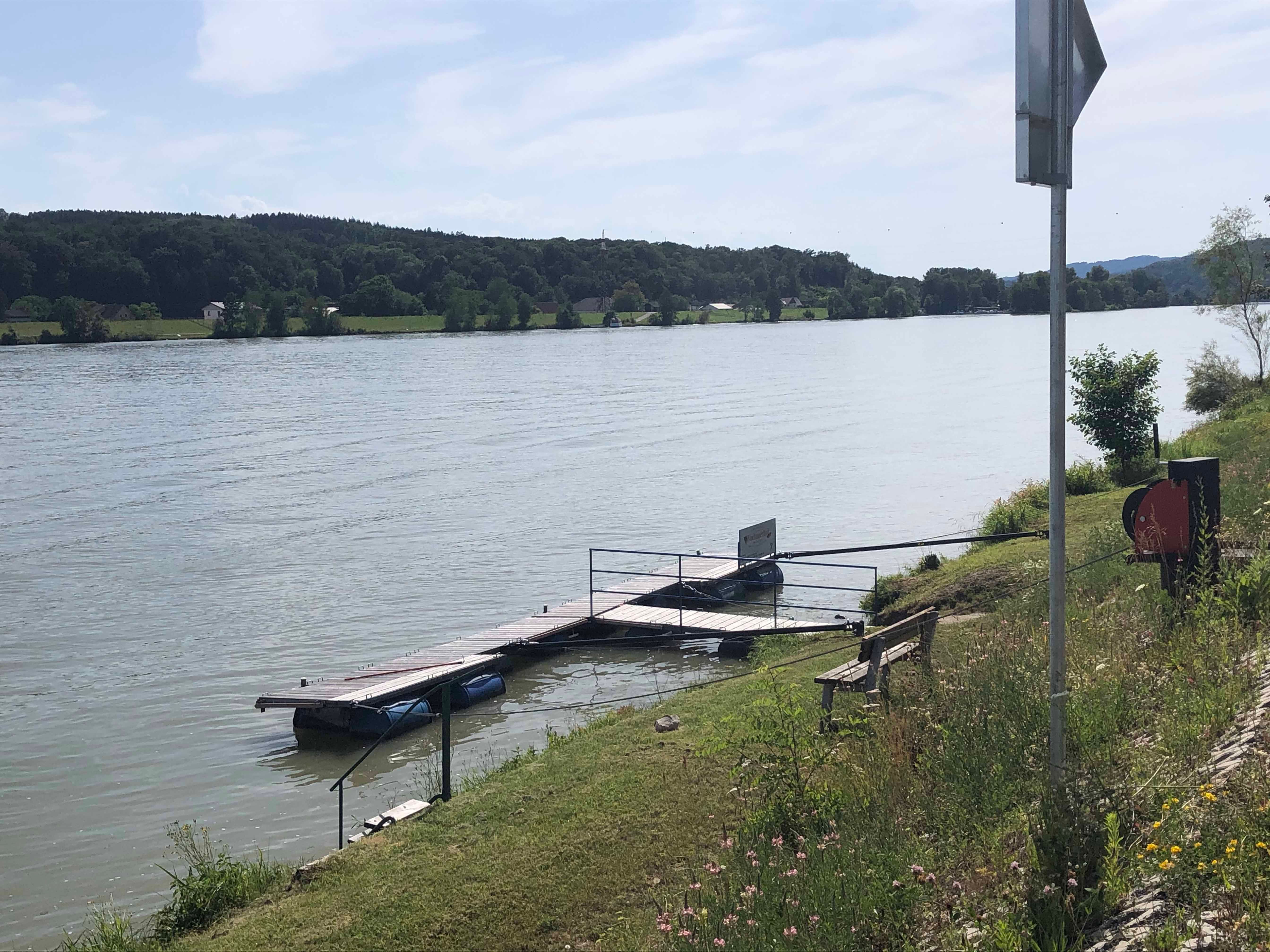 Mólo na brehu rieky s lavičkou a vegetáciou.