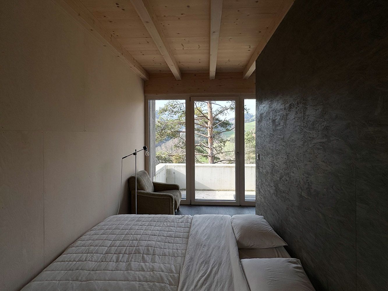 Minimalistická spálňa s posteľou, kreslom a oknom s výhľadom na stromy.