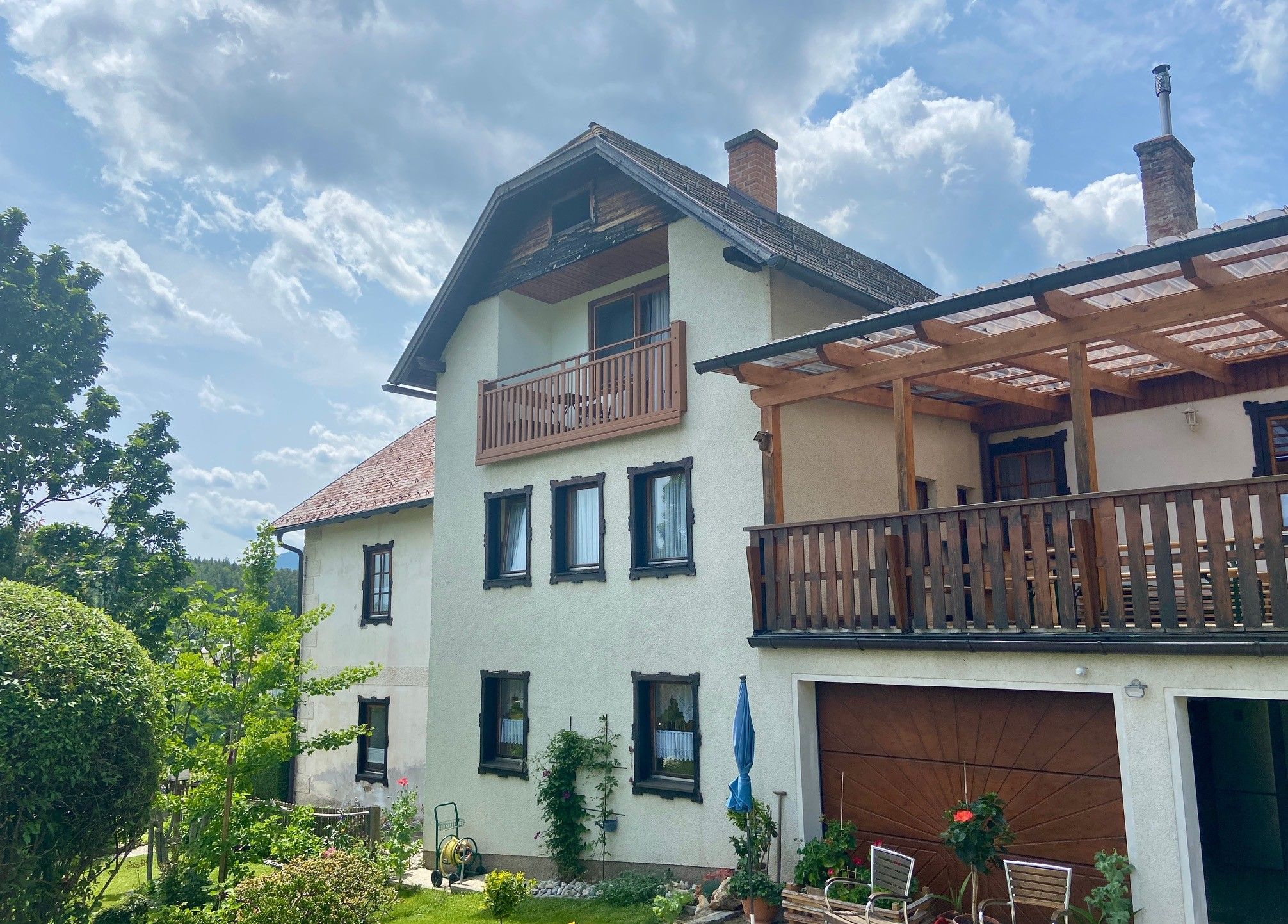 Dvojpodlažný dom s balkónom a terasou, obklopený záhradou a stromami, pod zamračenou oblohou.