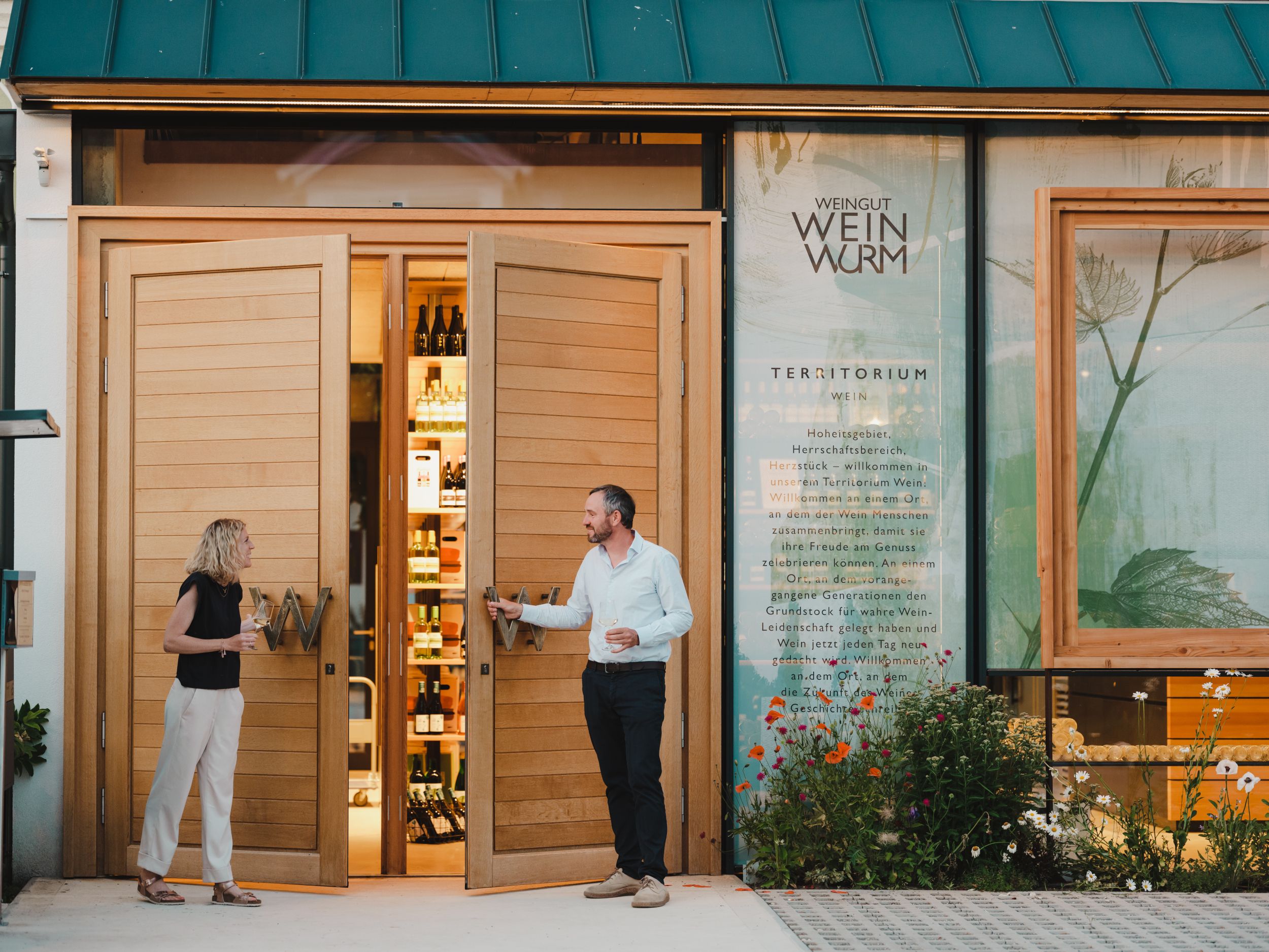 Dvaja ľudia stoja pred vinárňou s otvorenými drevenými dverami a fľašami vo vnútri.