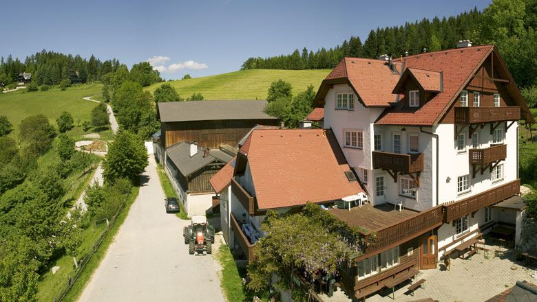 Farma s červenými strechami vo vidieckom prostredí, obklopená zelenými lúkami a lesmi.