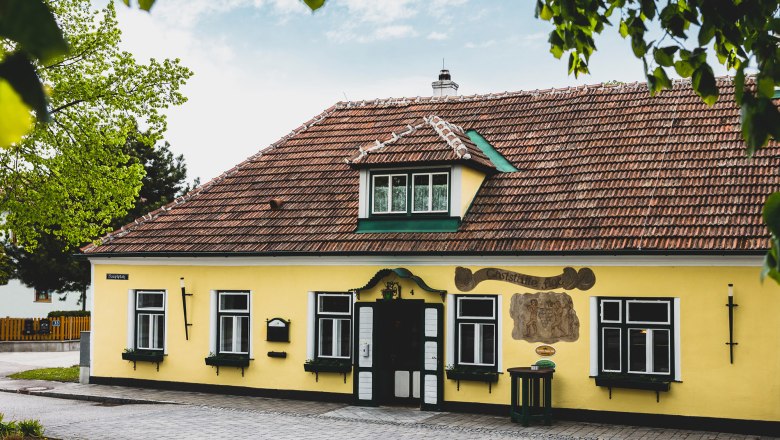 Žltá budova s červenými škridlami a zelenými okenicami, obklopená stromami.