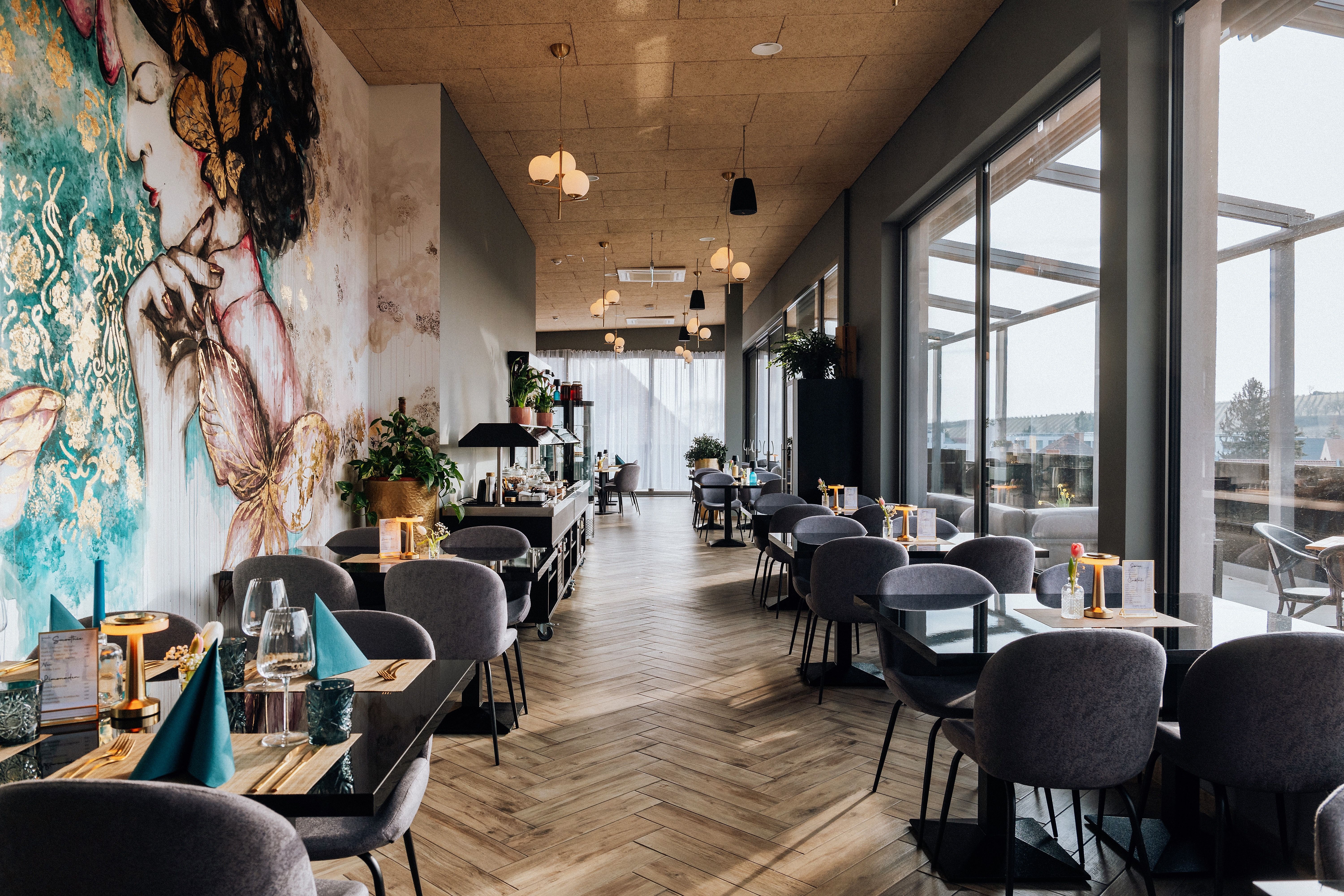 Pohľad do interiéru štýlovej reštaurácie s veľkými oknami a umeleckými nástennými maľbami.