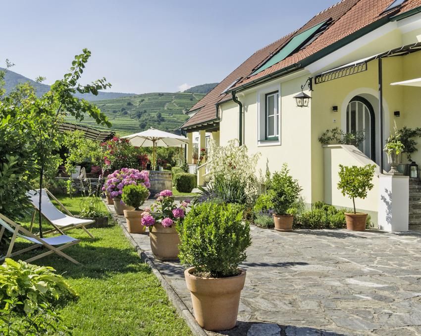 Idylický vidiecky dom s udržiavanou záhradou, kvitnúcimi rastlinami a ležadlami pod slnečnou oblohou.