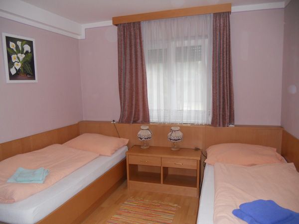 Jednoducho zariadená izba s dvoma samostatnými posteľami, oknom so závesmi a malým stolíkom s dvoma lampami.