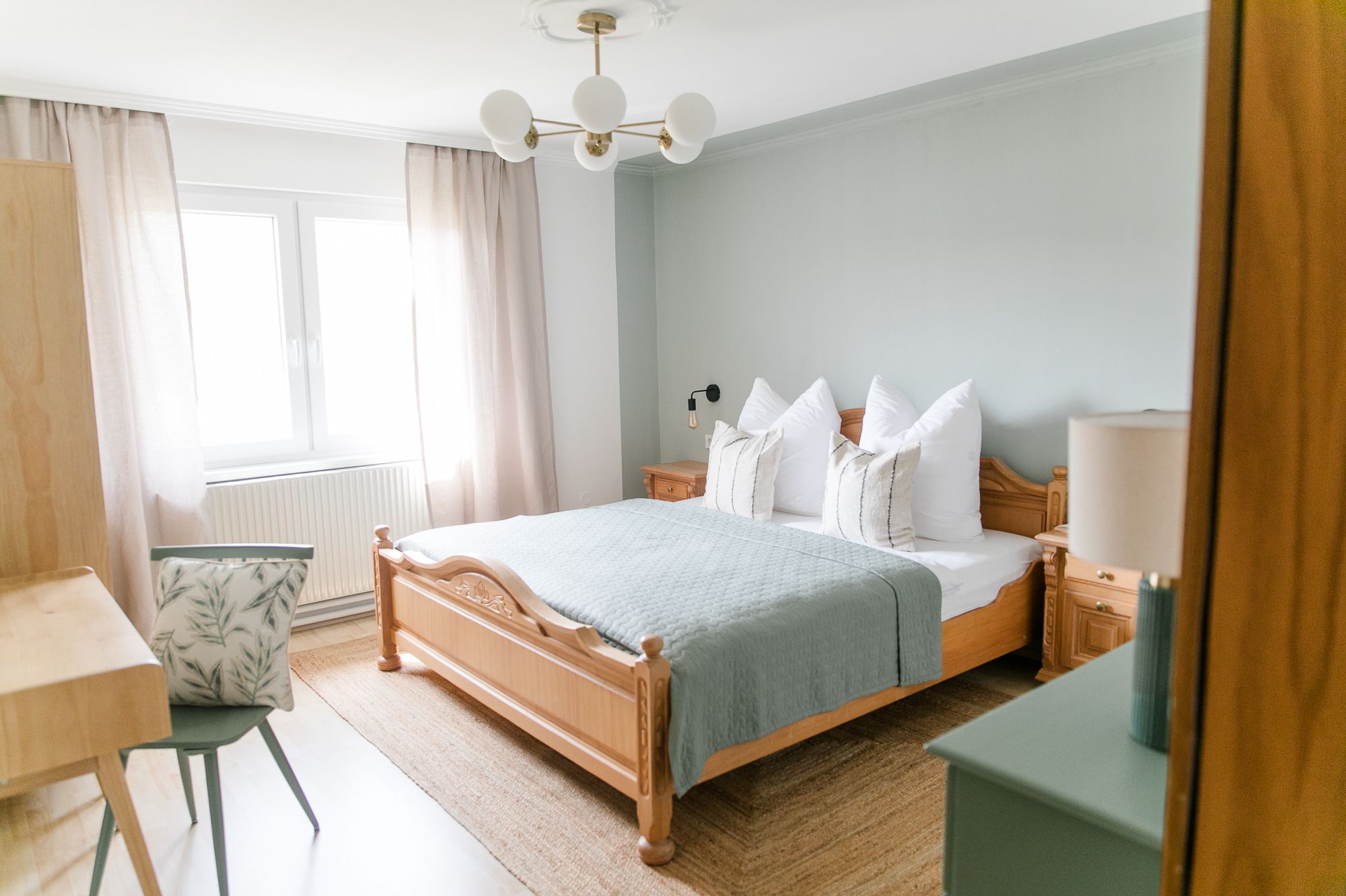 Útulná spálňa s drevenou posteľou, modrou perinou, nočnými stolíkmi a písacím stolom so stoličkou. Vidno svetlé závesy a okno.