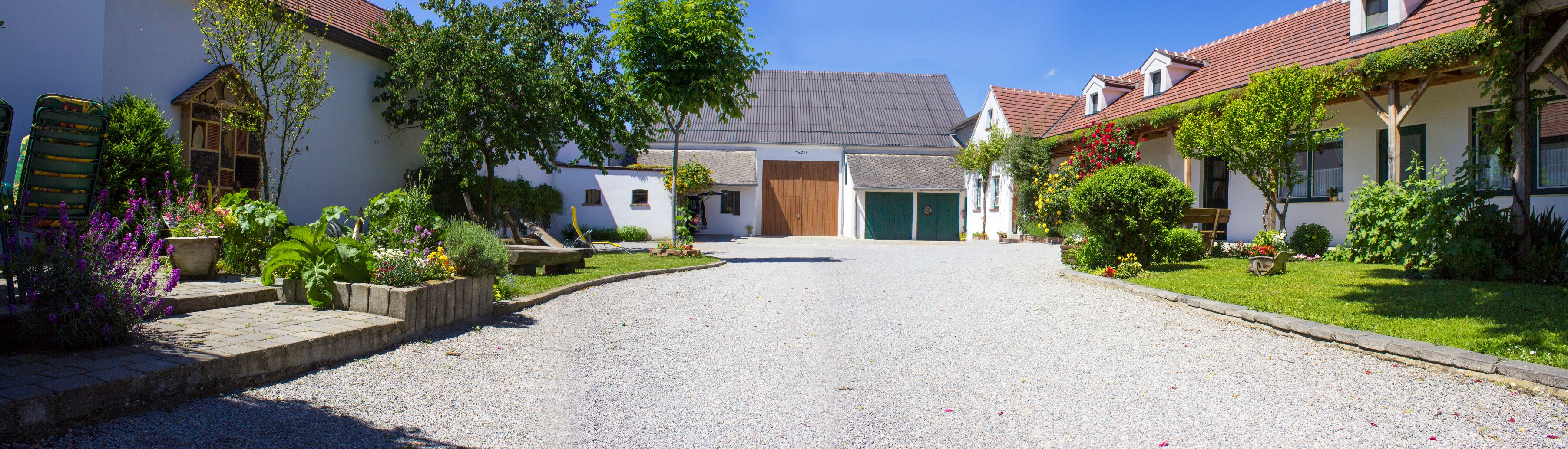 Idylická vinica so štrkovou cestou, kvitnúcimi záhradami a tradičnými budovami pod modrou oblohou.