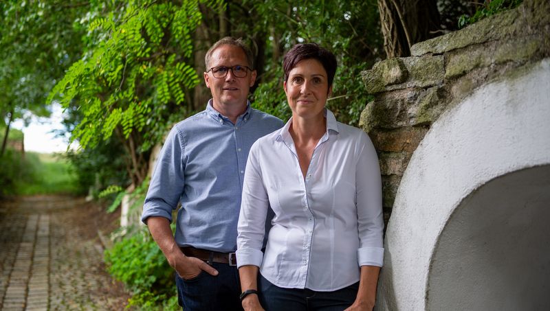Muž a žena stoja vedľa seba na dláždenej ceste pod holým nebom, obklopení stromami a kamenným múrom.