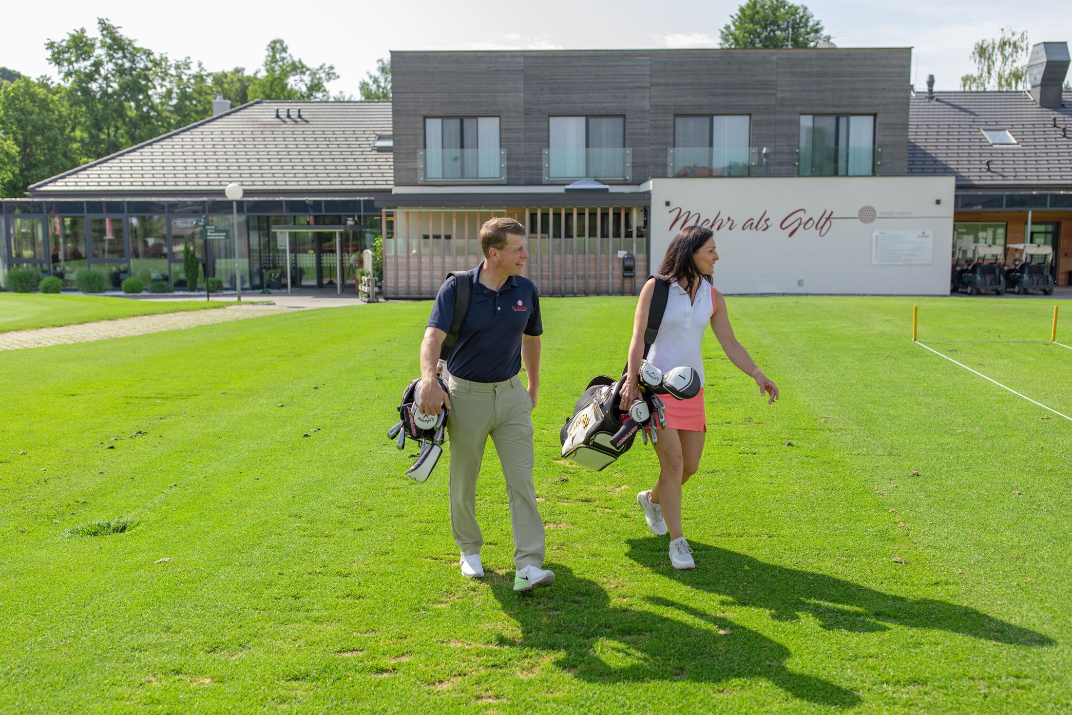 Dvaja ľudia s golfovými vakmi pred modernou klubovňou.