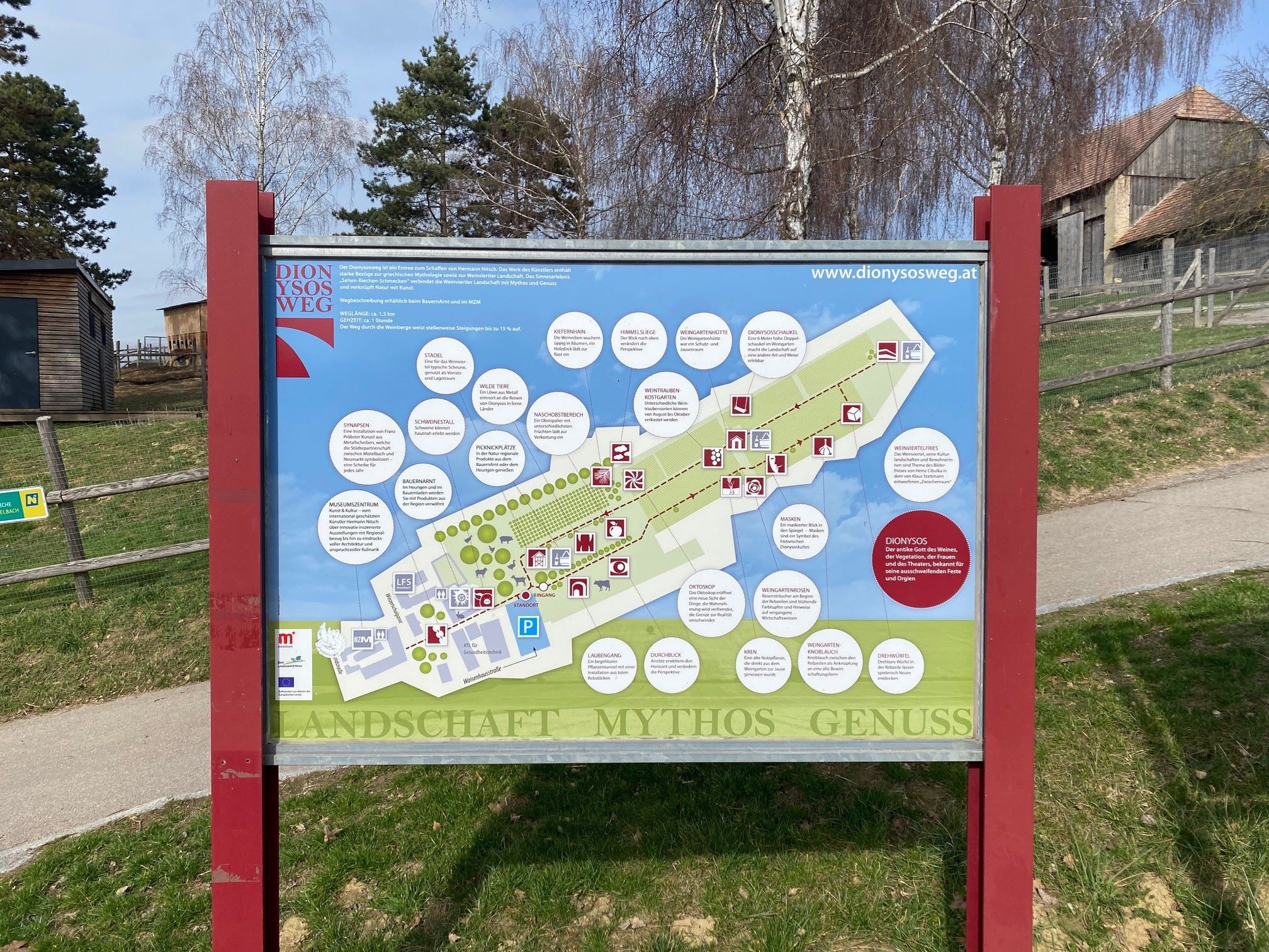 Informačná tabuľa Dionýzovej cesty s mapou a vysvetlivkami, obklopená stromami a budovami.