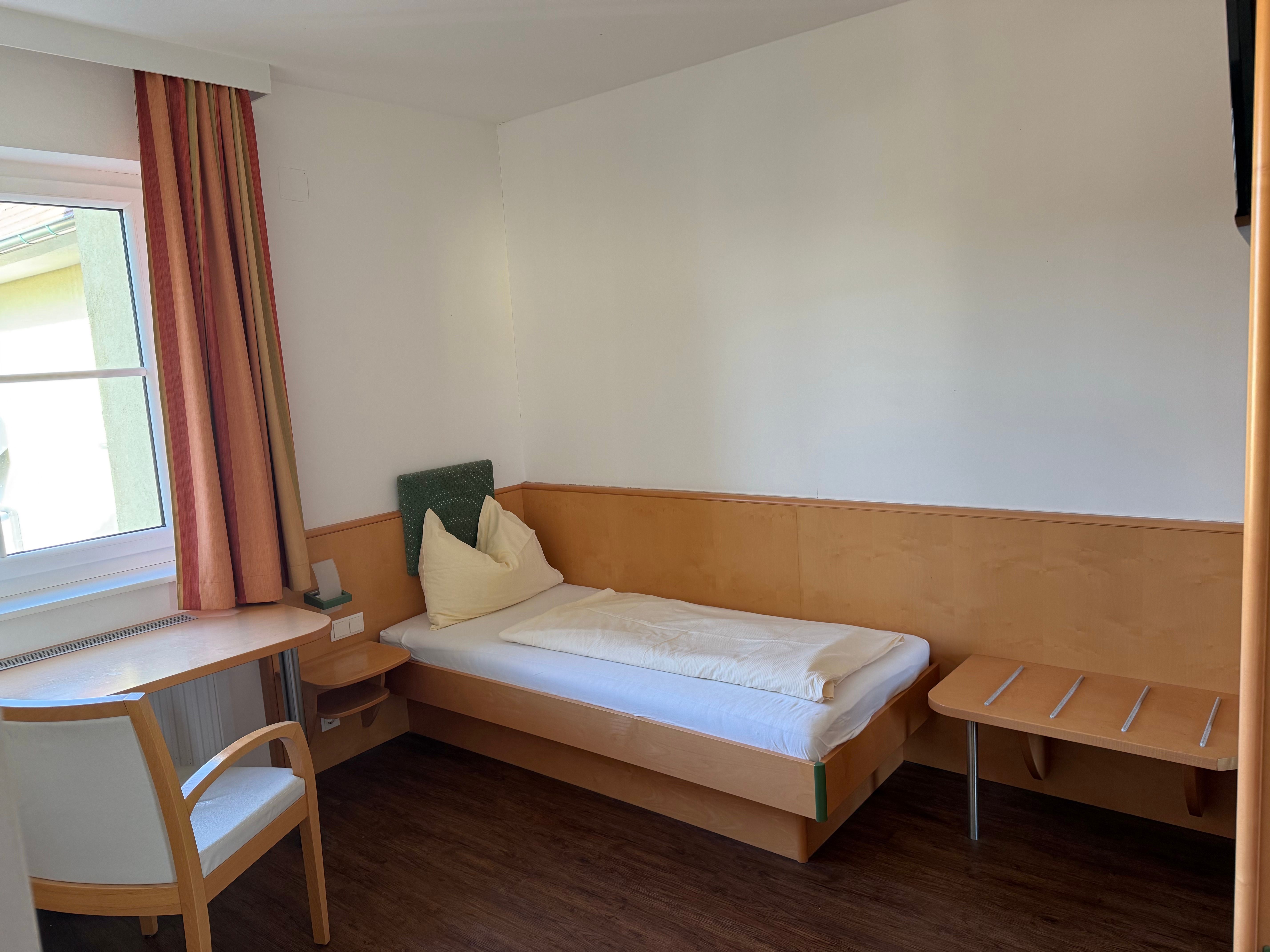 Jednoduchá hotelová izba s jednou posteľou, stolom, stoličkou a oknom so závesmi.