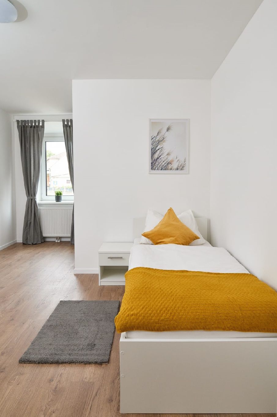 Jednolôžková izba s posteľou, nočným stolíkom a oknom so závesmi.