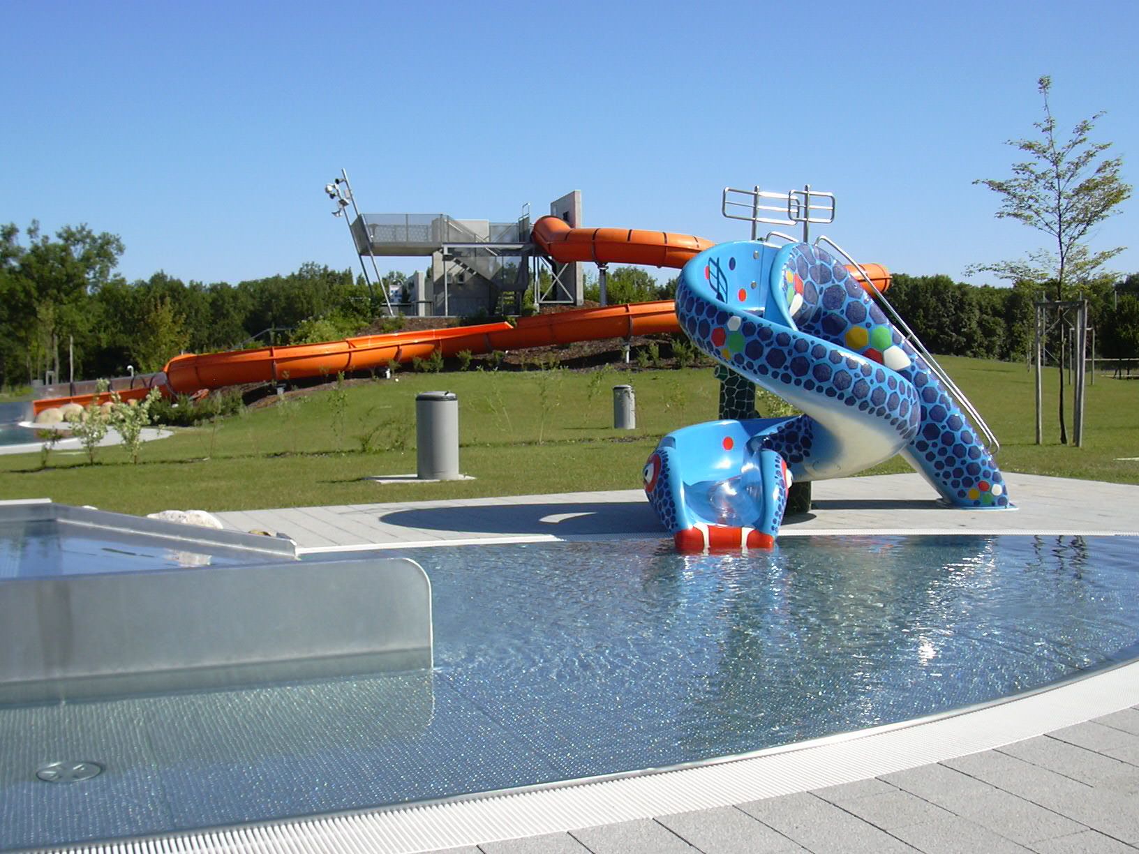 Pohľad na aquapark s modrou detskou šmýkačkou a veľkým oranžovým toboganom v pozadí.