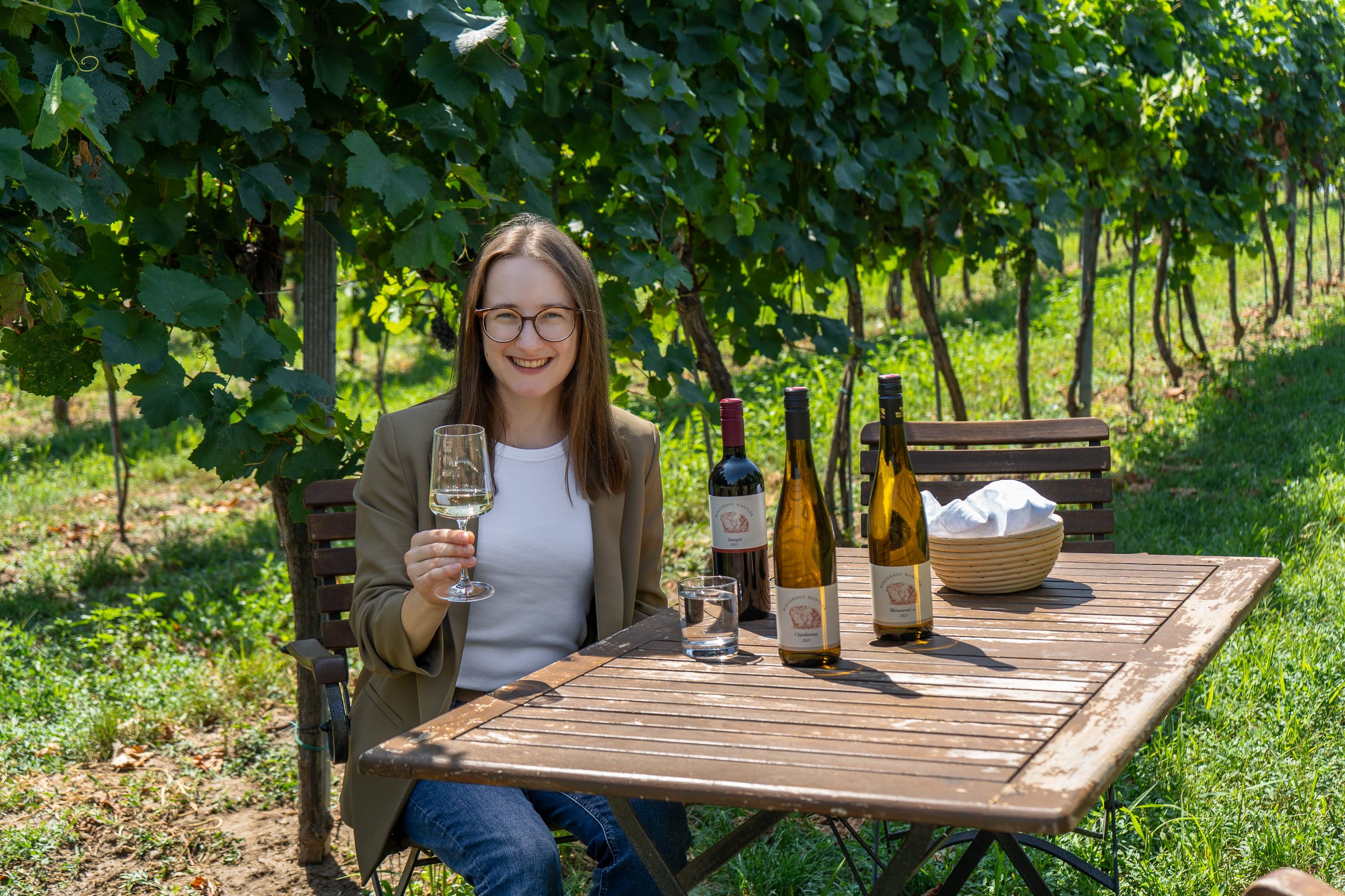 Žena sedí za dreveným stolom vo vinici a v ruke drží pohár s vínom. Na stole je niekoľko fliaš vína.