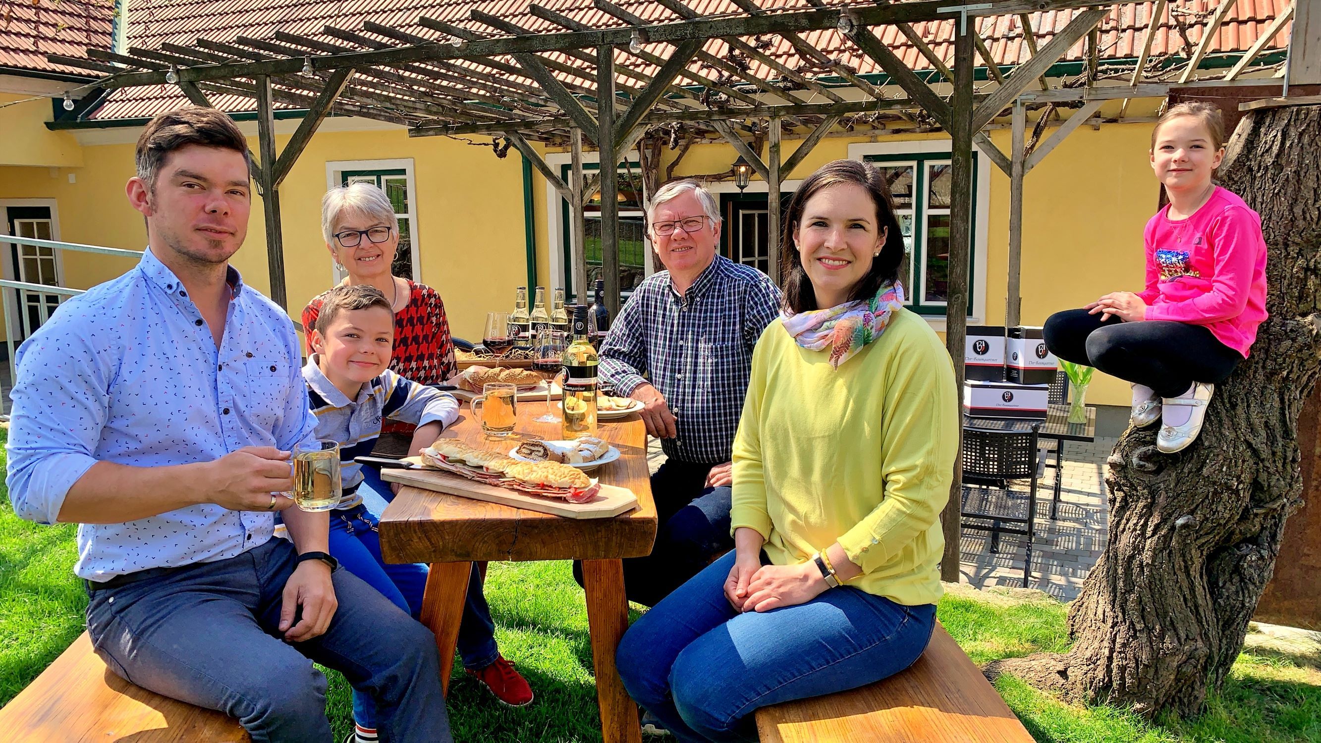 Rodina sedí vonku pri drevenom stole a vychutnáva si jedlo. Dieťa sedí na kmeni stromu. V pozadí je žltý dom s pergolou.