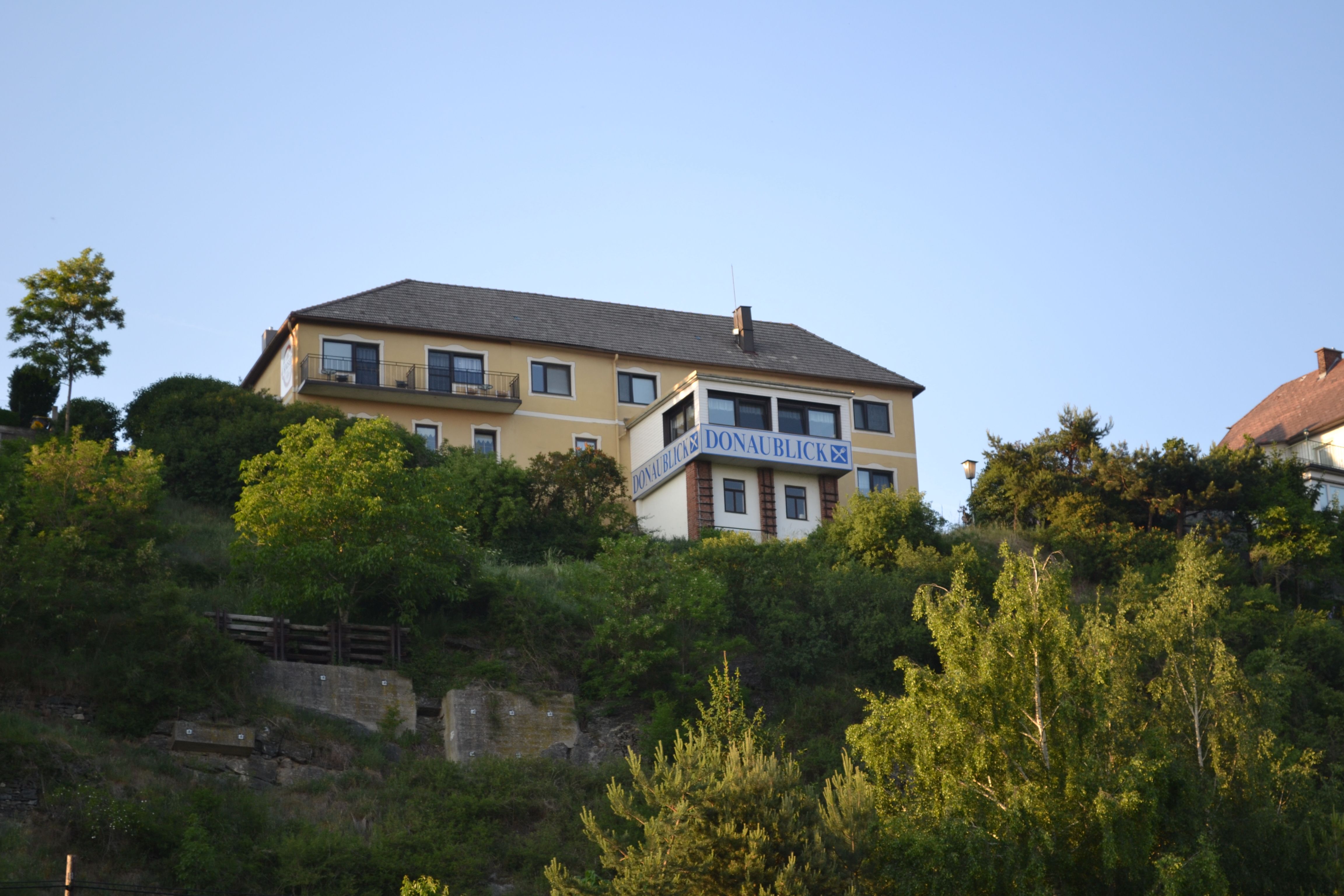Žltá budova s nápisom "Donaublick" na kopci, obklopená stromami a modrou oblohou.