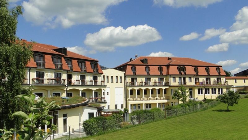 Kurhotel Moorbad Großpertholz s červenou strechou a balkónmi, obklopený zelenými lúkami a stromami pod modrou oblohou.