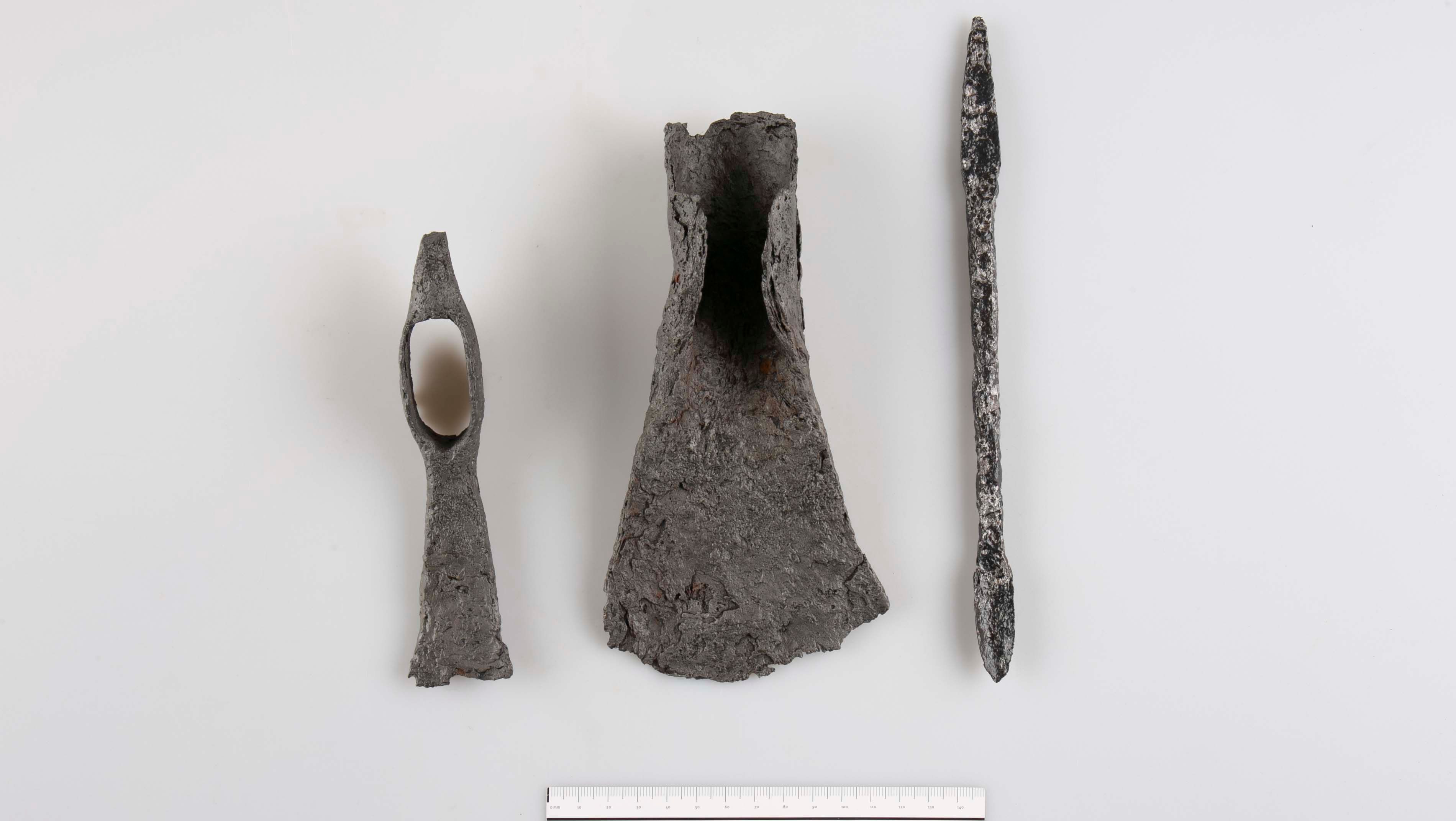 Tri archeologické kovové predmety na bielom pozadí so stupnicou v spodnej časti.