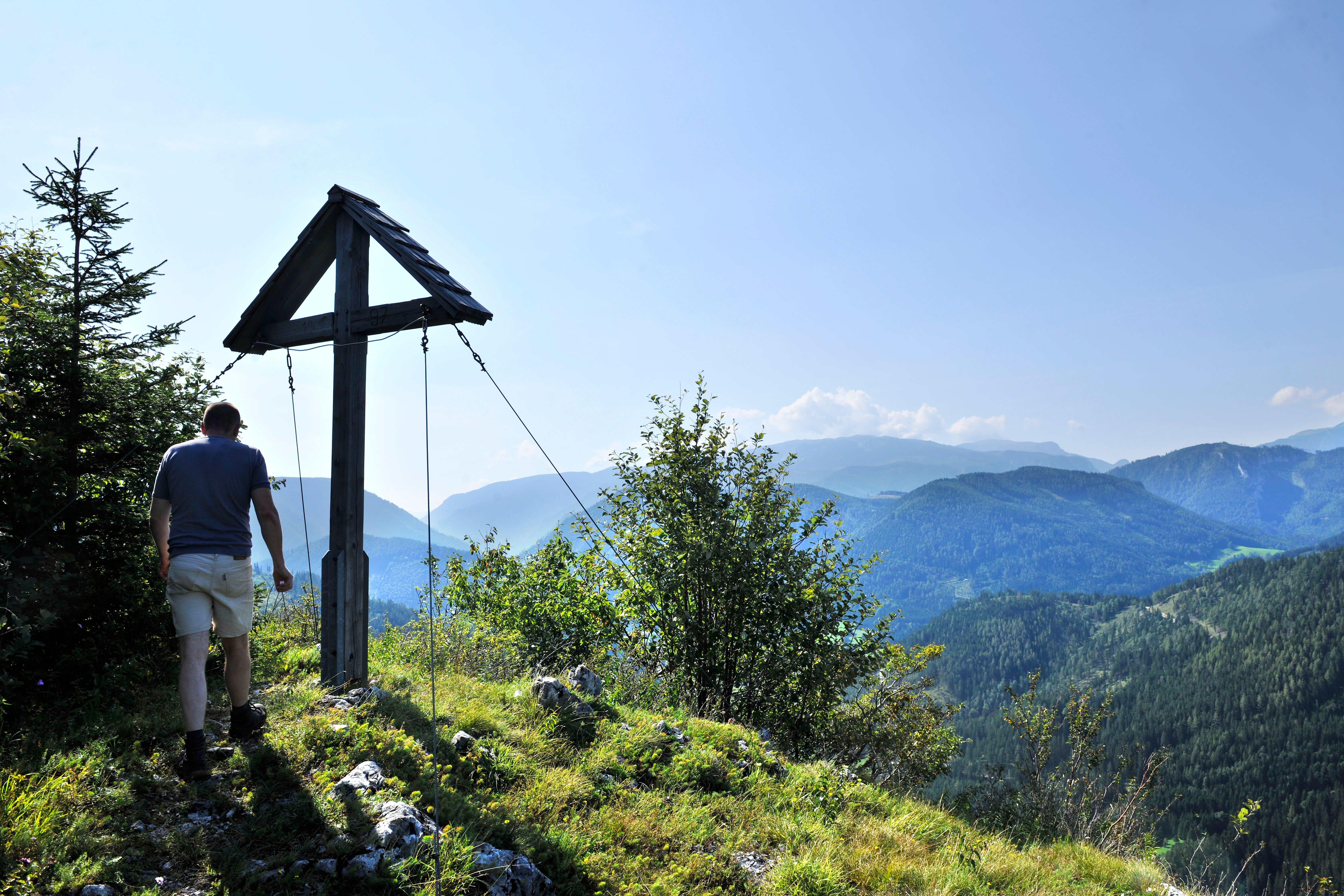 Turista stojí pri vrcholovom kríži s výhľadom na zalesnené hory.