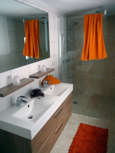 Moderná kúpeľňa s dvojitým umývadlom, veľkým zrkadlom, sprchovacím kútom a oranžovými akcentmi.