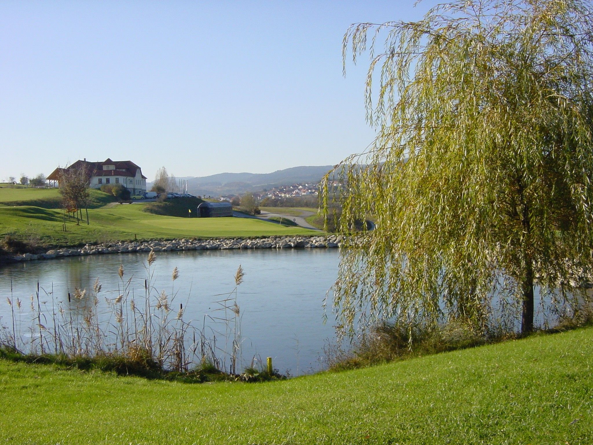 Golfové ihrisko s rybníkom, stromami a budovou v pozadí.