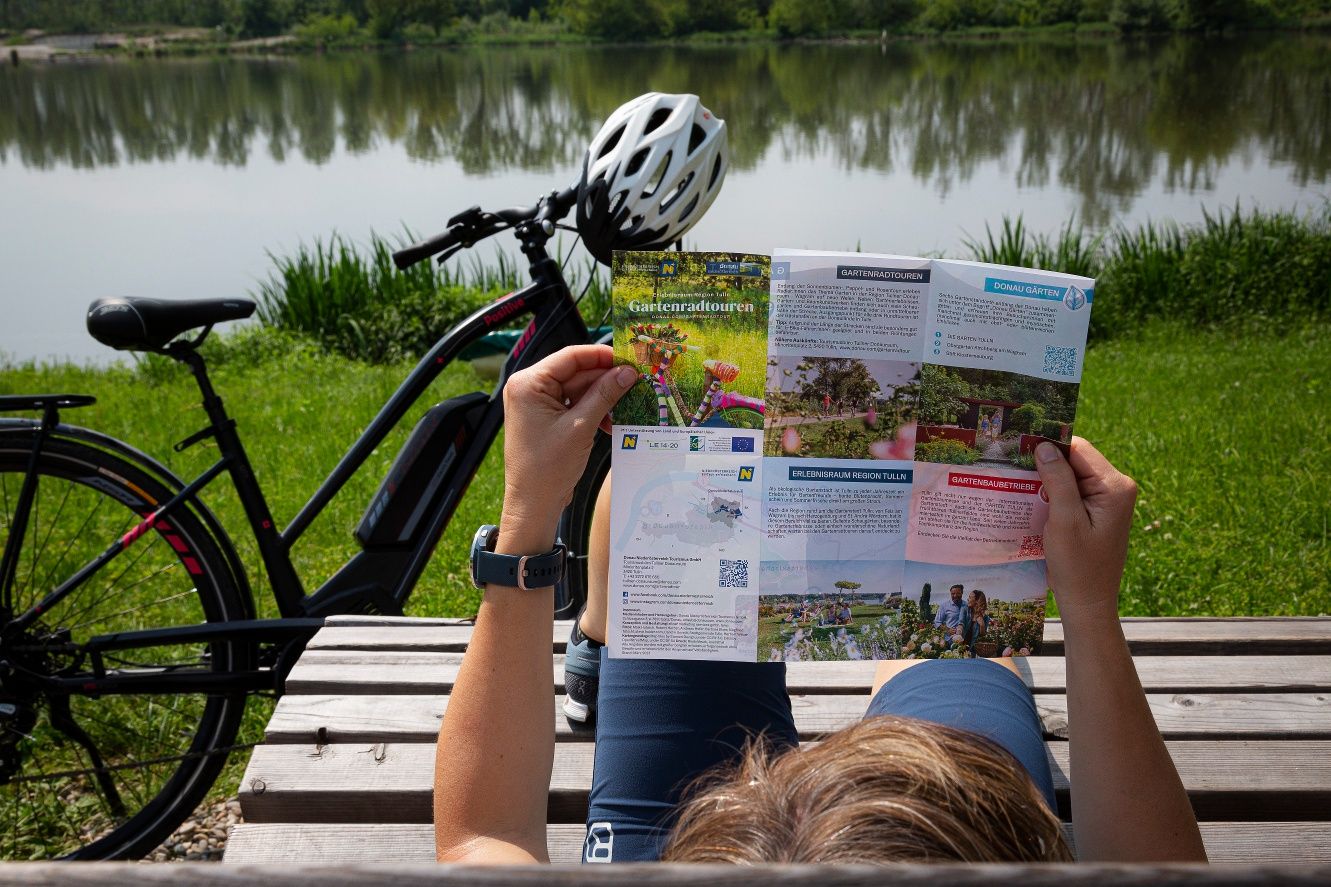 Osoba si číta skladaciu mapu o záhradných cyklotúrach vedľa bicykla pri jazere.